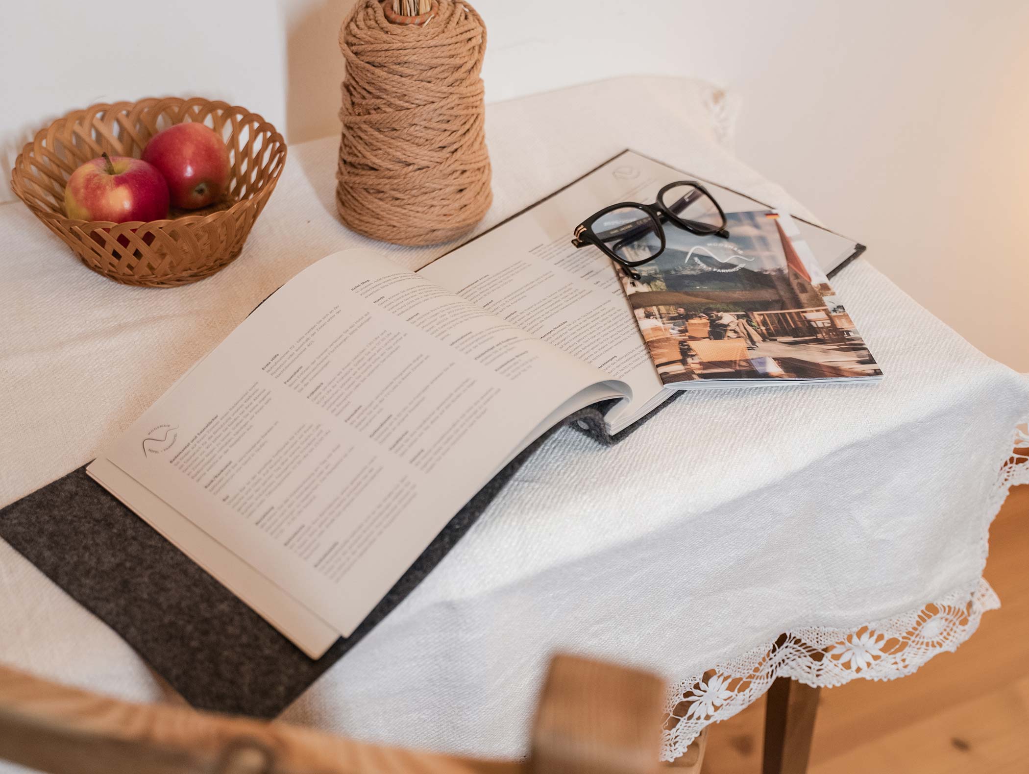 Ein aufgeschlagenes Buch, Gläser und eine Zeitschrift auf einem Tisch mit Äpfeln und Schnüren, bedeckt mit einem Spitzentuch. - Hotel Moosmair