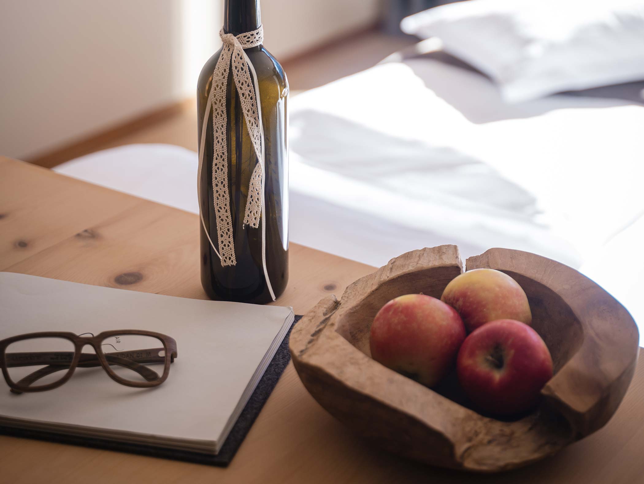 Eine Flasche mit einem Spitzenband, Äpfel in einer Holzschale, Gläser und ein Notizbuch auf einem Holztisch. - Hotel Moosmair
