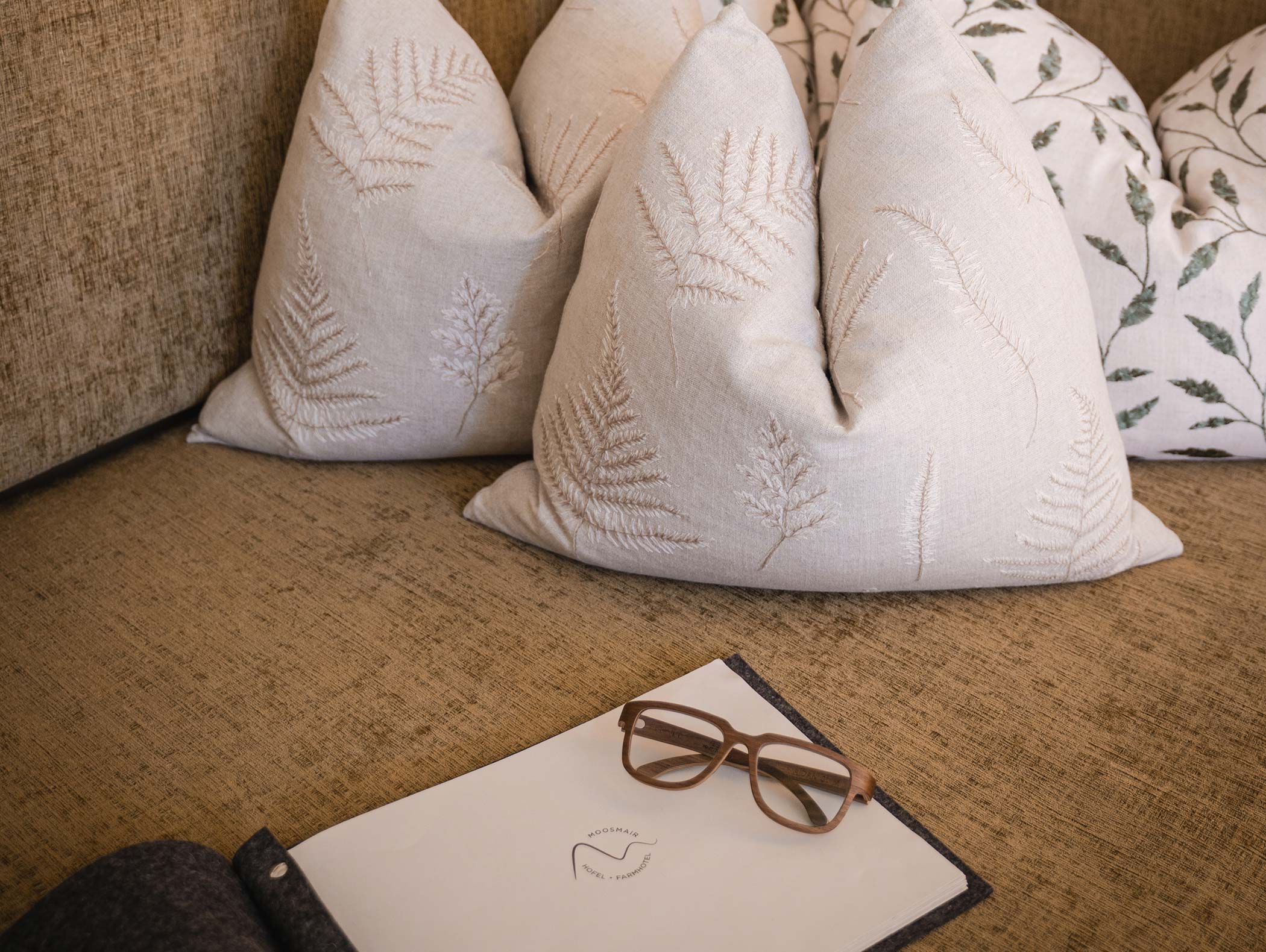 Braune Couch mit farngemusterten Kissen, Brille und einem geschlossenen Notizbuch auf dem Sitz. - Hotel Moosmair