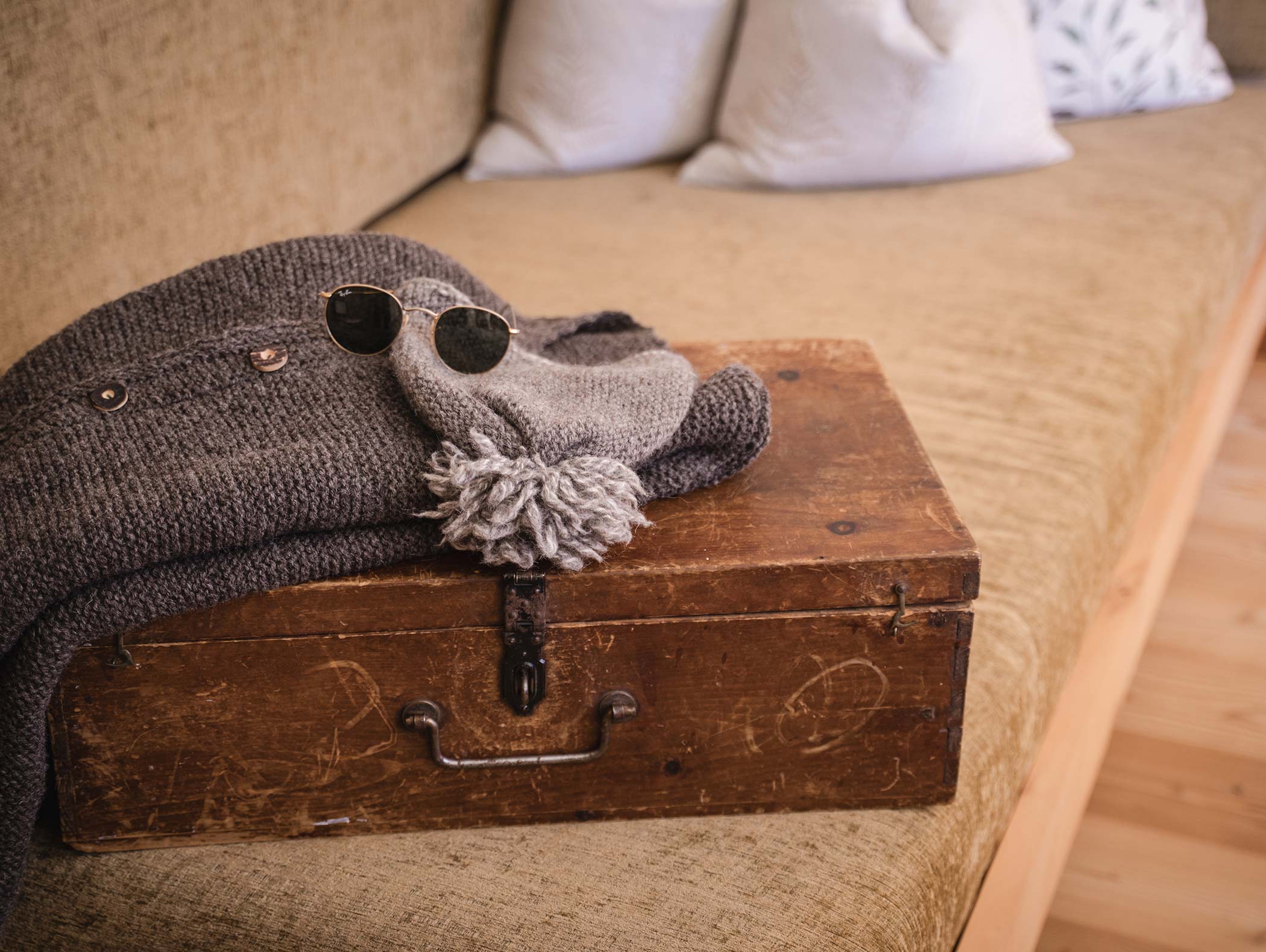 Ein Strickpullover, eine graue Mütze und eine Sonnenbrille liegen auf einem alten Koffer auf einer beigen Couch. - Hotel Moosmair