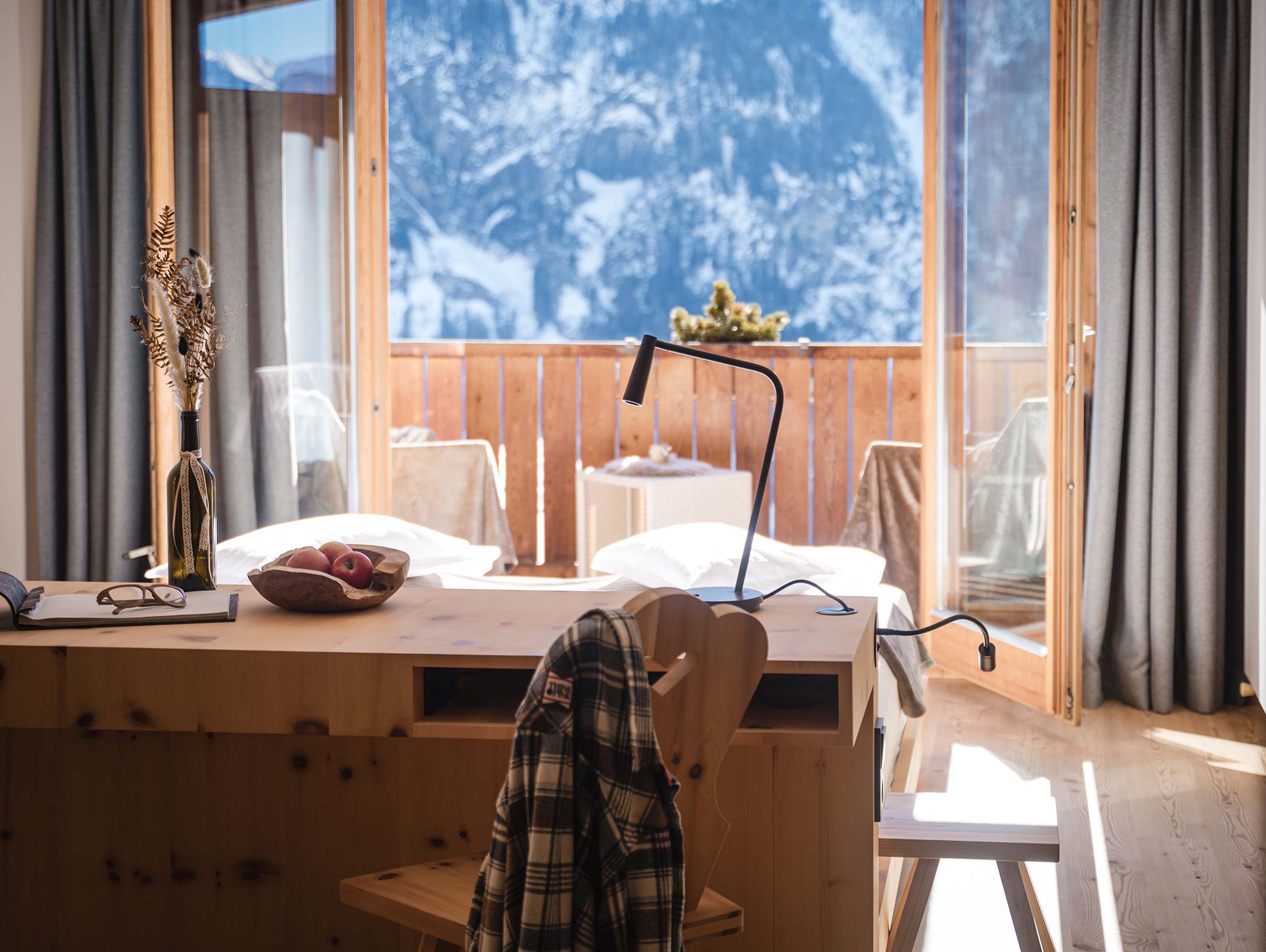 Ein gemütliches Holzzimmer mit Schreibtisch, Bett und Blick auf die Berge durch große Glastüren auf einen sonnenbeschienenen Balkon. - Hotel Moosmair