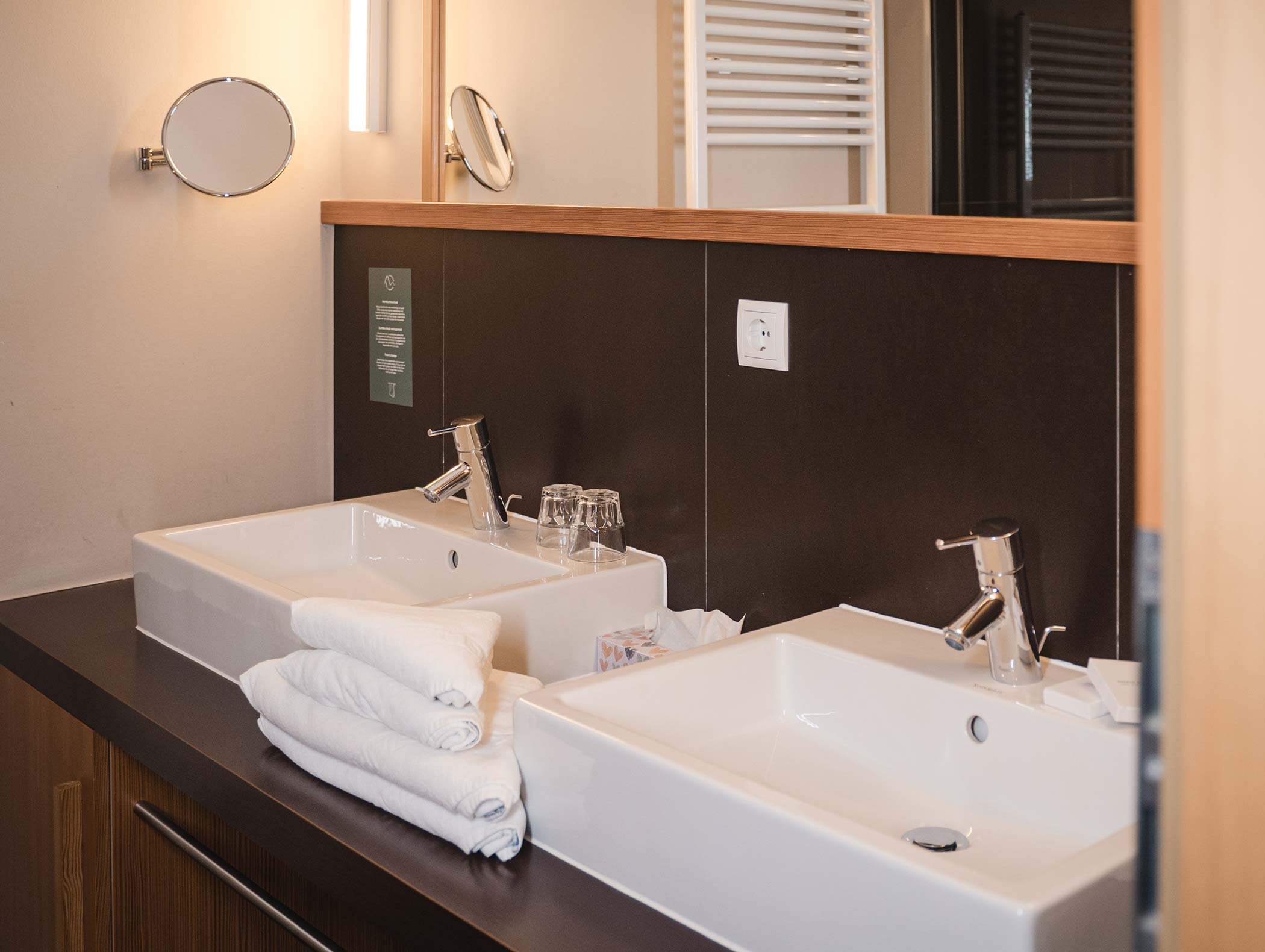 Zwei weiße Waschbecken mit verchromten Armaturen, Handtücher und Gläser auf einer dunklen Arbeitsplatte in einem modernen Badezimmer. - Hotel Moosmair