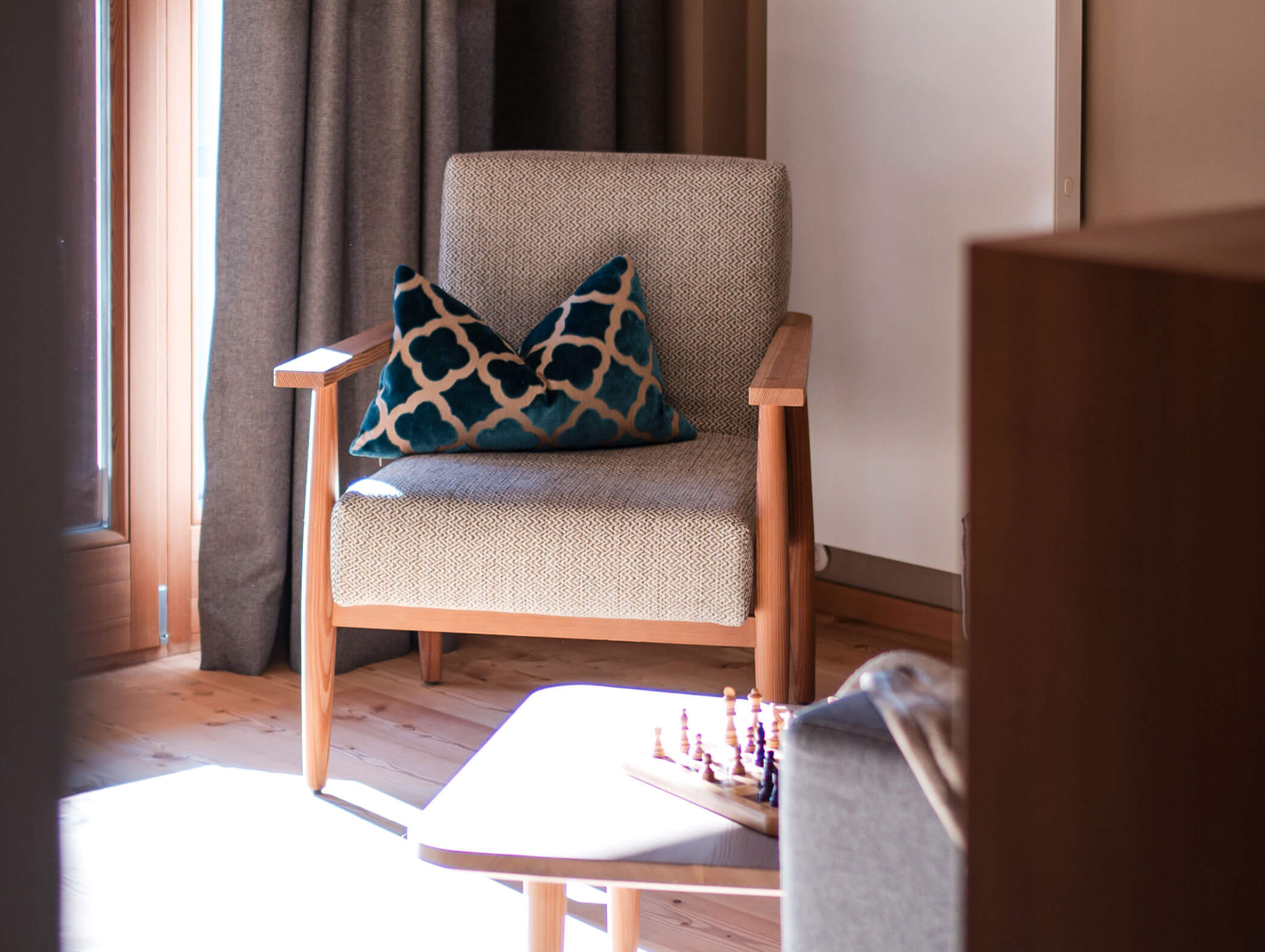 Beigefarbener Sessel mit einem blau gemusterten Kissen neben einem sonnenbeschienenen Tisch mit einem Schachspiel in einem gemütlichen Zimmer. - Hotel Moosmair