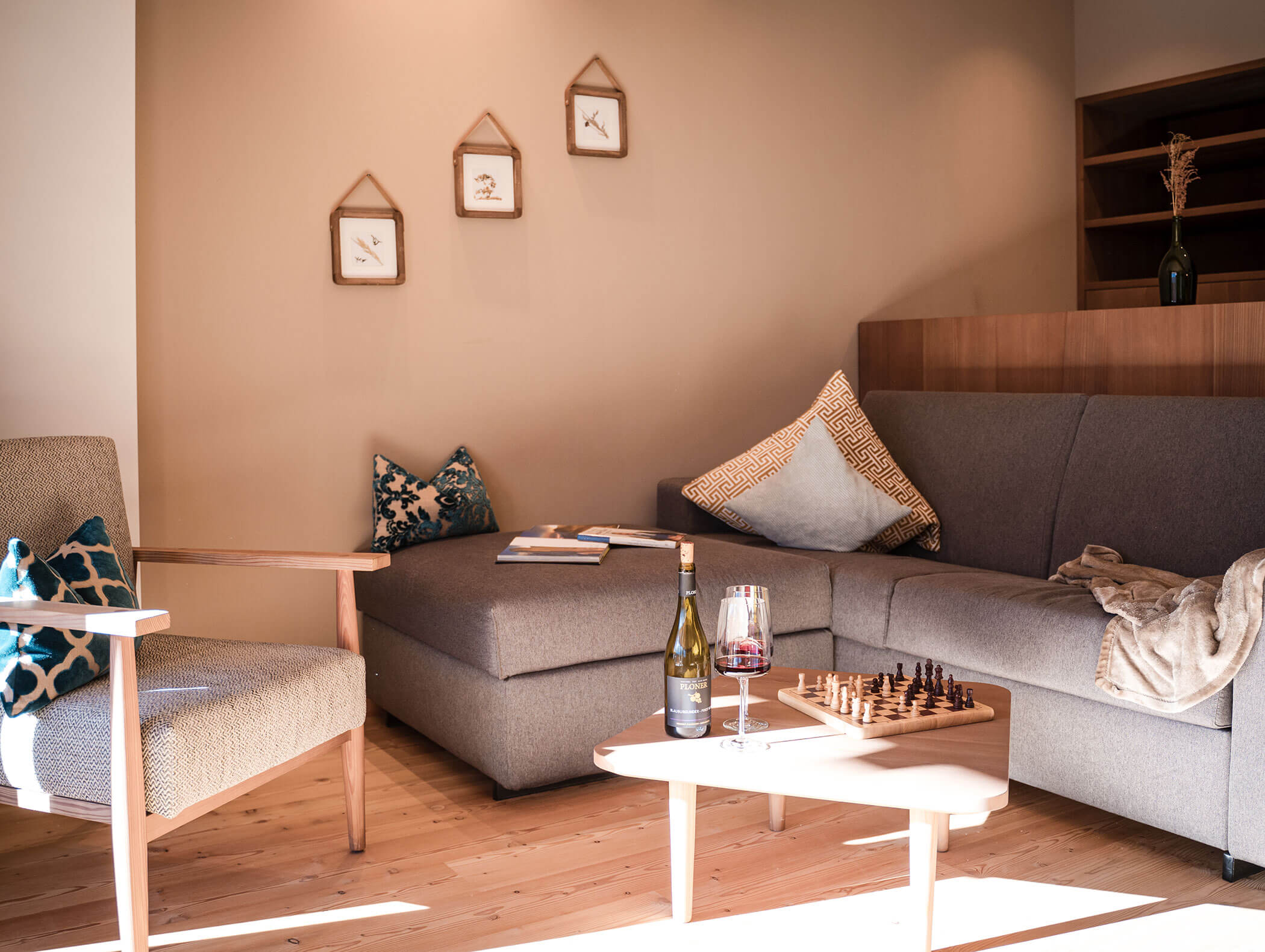 Gemütliches Wohnzimmer mit einem grauen Sofa, Sessel, Wein, Schachbrett und gerahmter Kunst an einer beigen Wand. - Hotel Moosmair