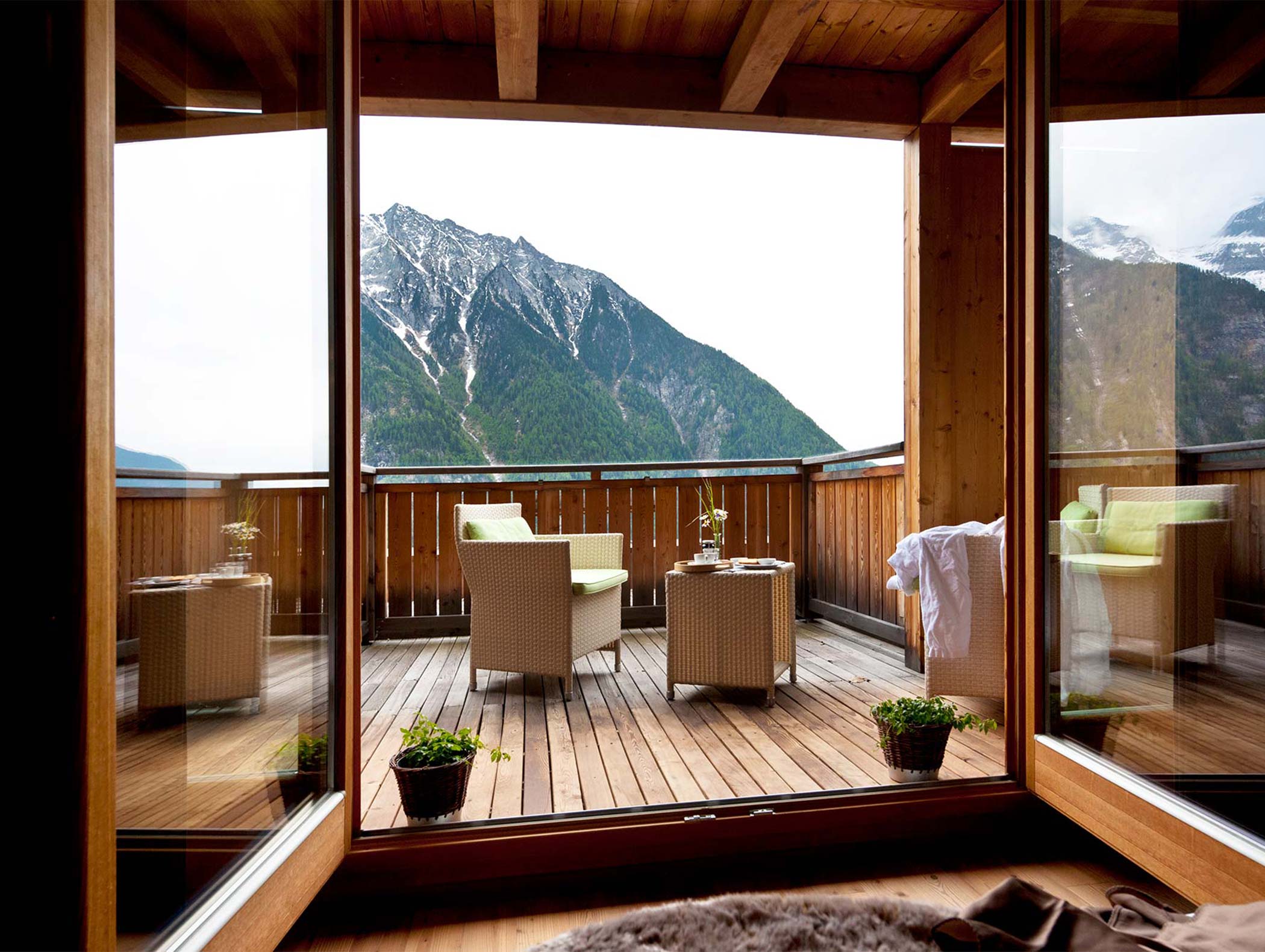 Blick auf die Berge von einer Holzterrasse mit Stühlen und kleinen Tischen, die durch große Glastüren zu sehen ist. - Hotel Moosmair