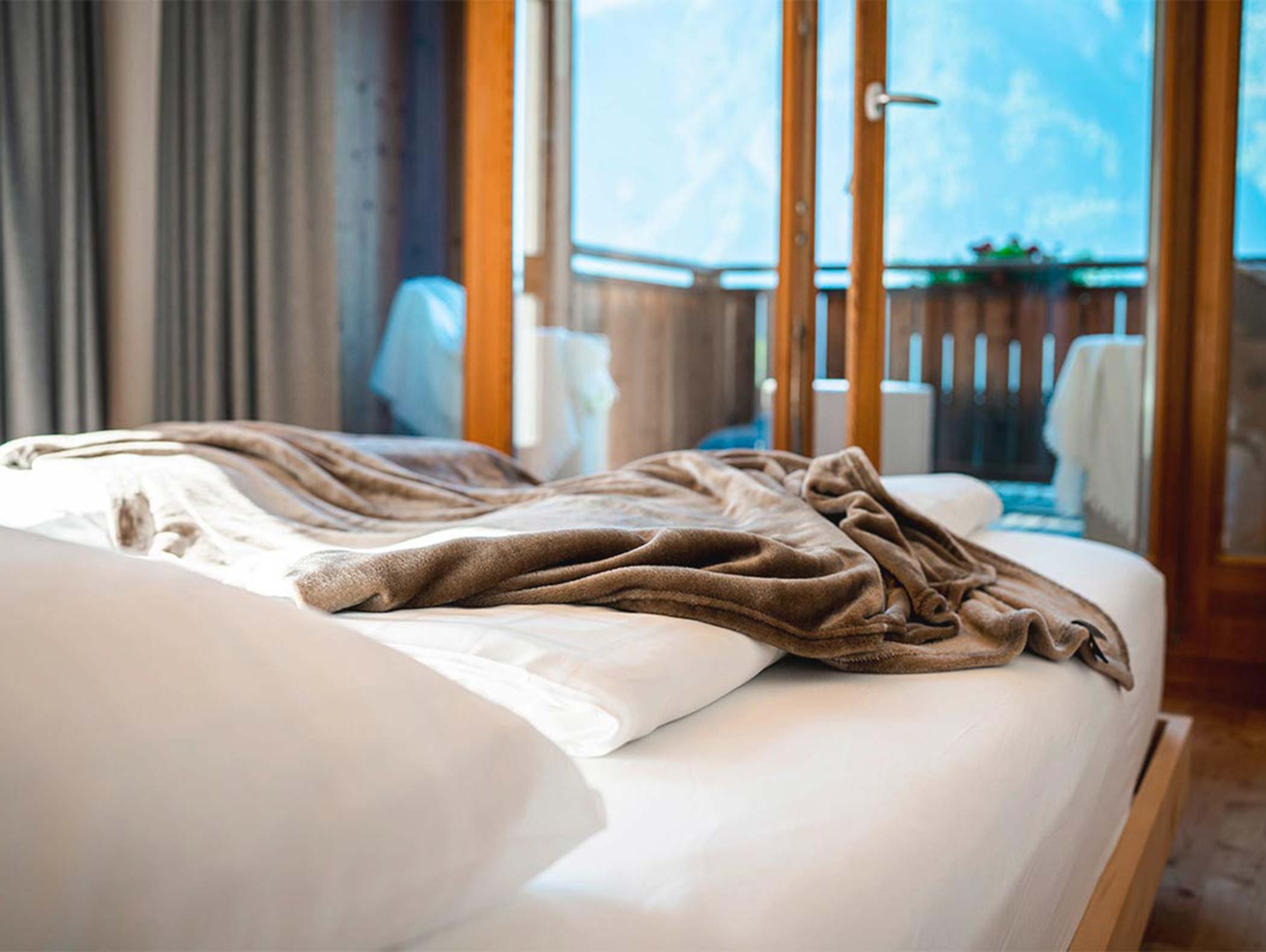 Ungemachtes Bett mit weißen Laken und einer braunen Decke in einem hellen Zimmer mit offenen Türen zu einem Balkon. - Hotel Moosmair