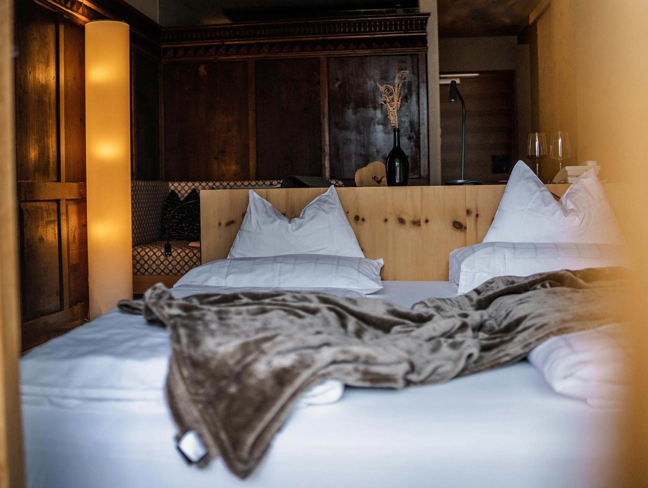 Gemütliches Schlafzimmer mit weißen Kissen, einer braunen Decke und Holzdekor, sanft beleuchtet von einer hohen Stehlampe. - Hotel Moosmair