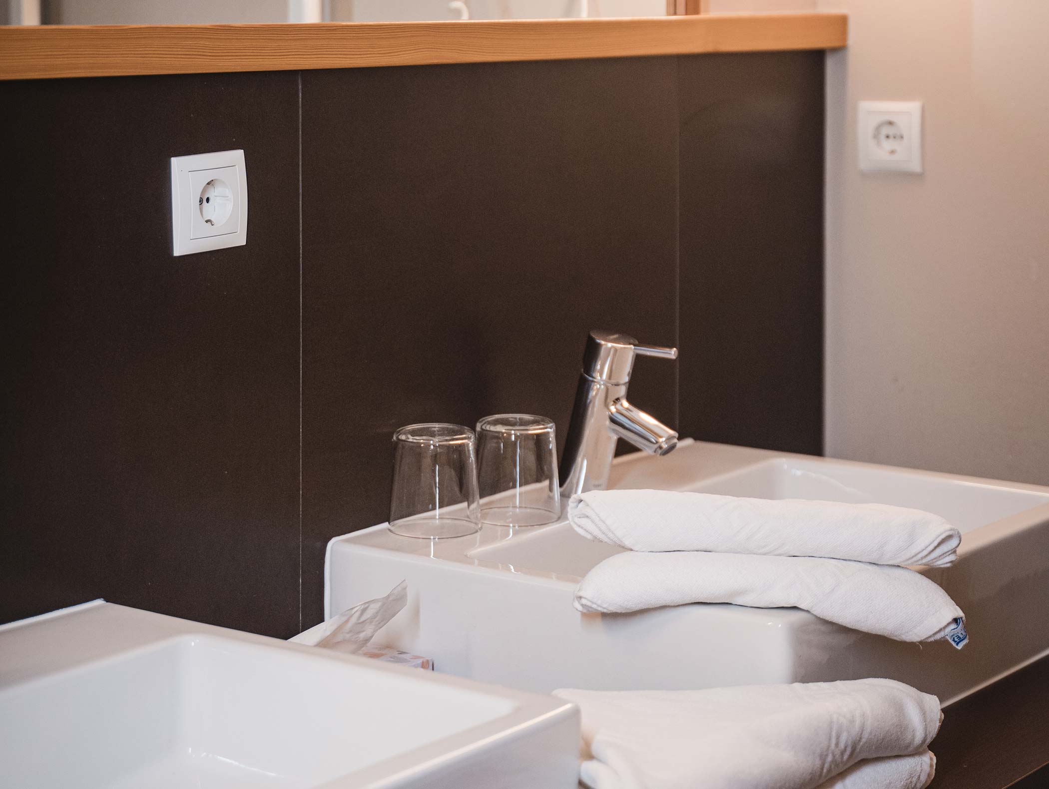 Ein modernes Waschbecken mit gefalteten weißen Handtüchern, zwei Gläsern und einem verchromten Wasserhahn in der Nähe einer dunklen Wand mit Steckdosen. - Hotel Moosmair