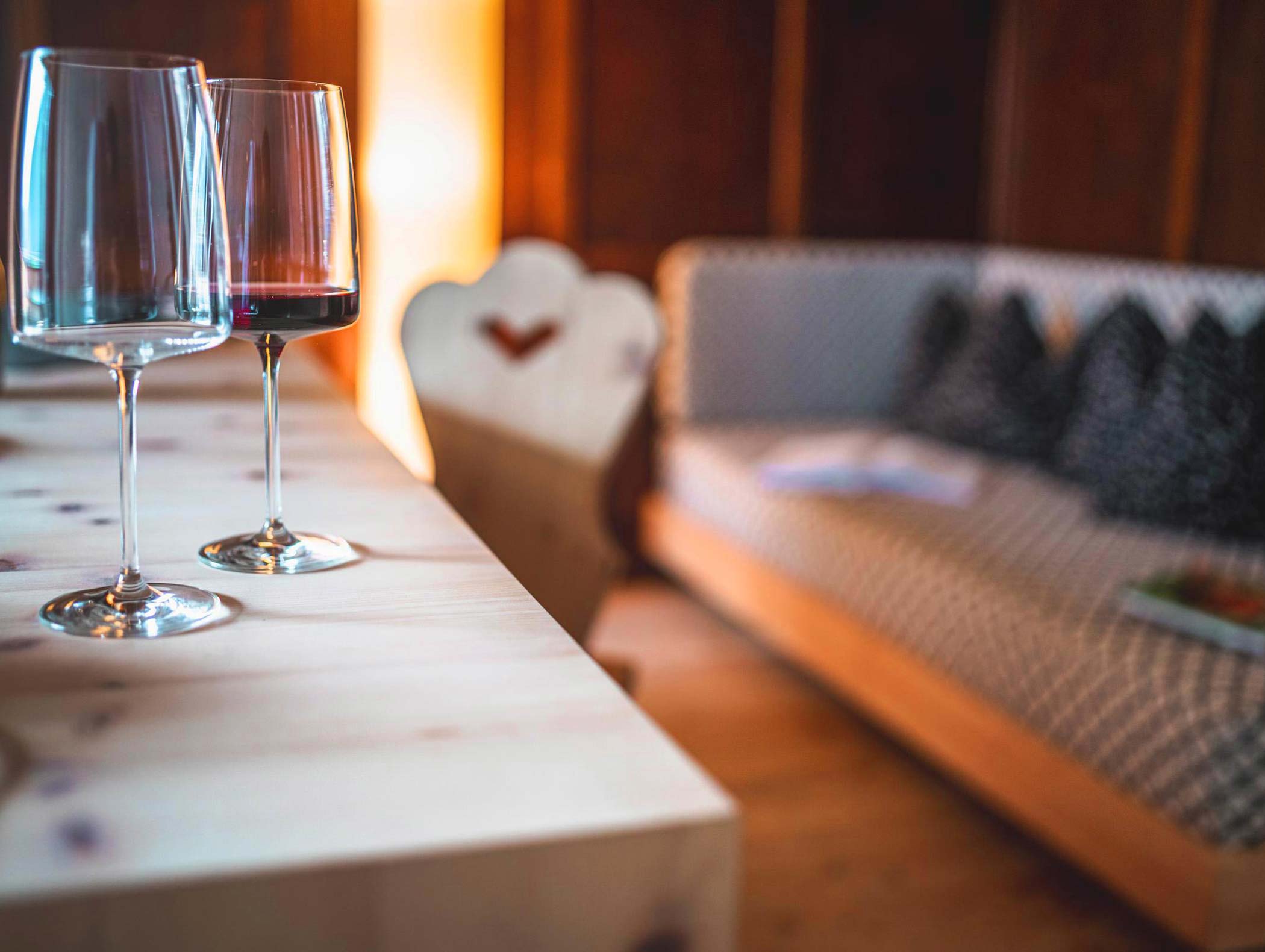Zwei Weingläser auf einem Holztisch mit einem verschwommenen Sofa und Sessel im Hintergrund. - Hotel Moosmair