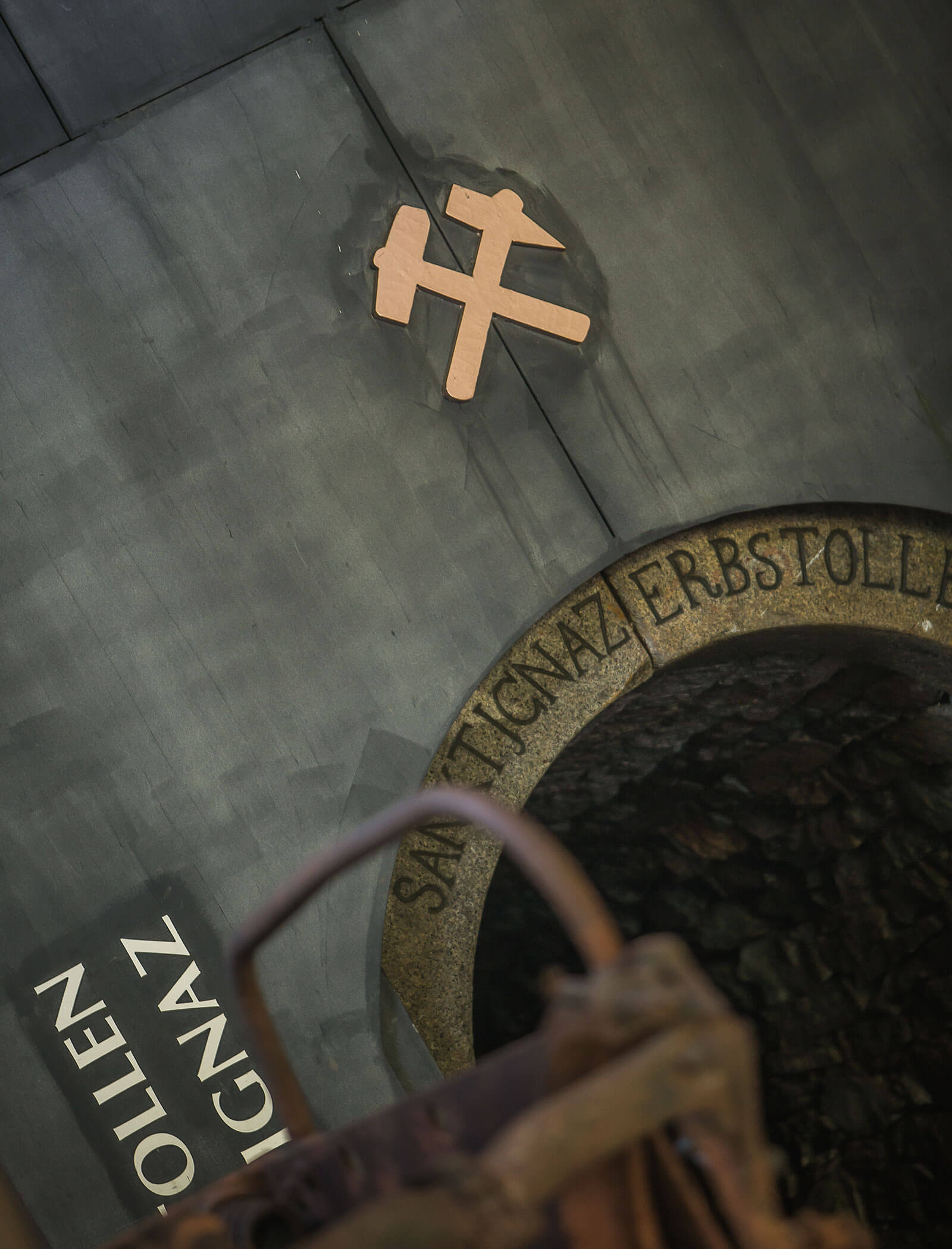Ein Bergbausymbol über einem dunklen steinernen Tunneleingang mit der Aufschrift SANKT IGNATZ ERBSTOLLEN. - Hotel Moosmair
