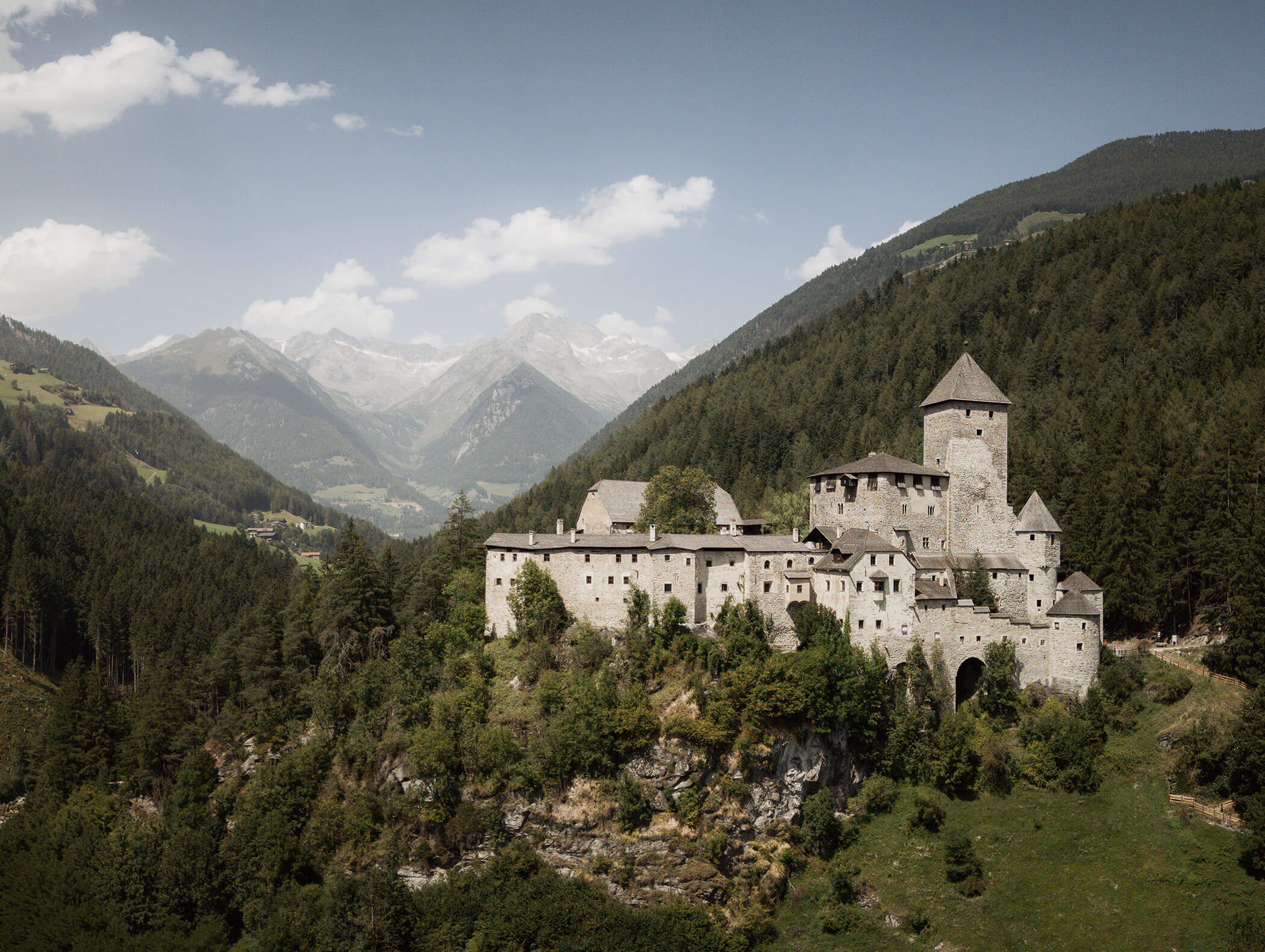 Die mittelalterliche Steinburg liegt auf einem bewaldeten Hügel mit Bergen im Hintergrund unter einem teilweise bewölkten Himmel. - Hotel Moosmair