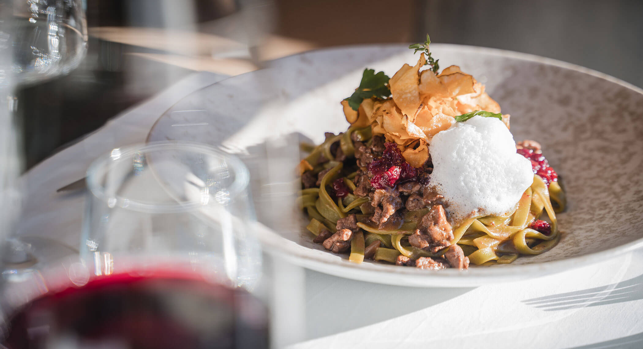 Ein Teller mit Gourmet-Pasta mit Fleisch, Schaum, knusprigen Beilagen und Kräutern auf einem weißen Tischtuch. - Hotel Moosmair