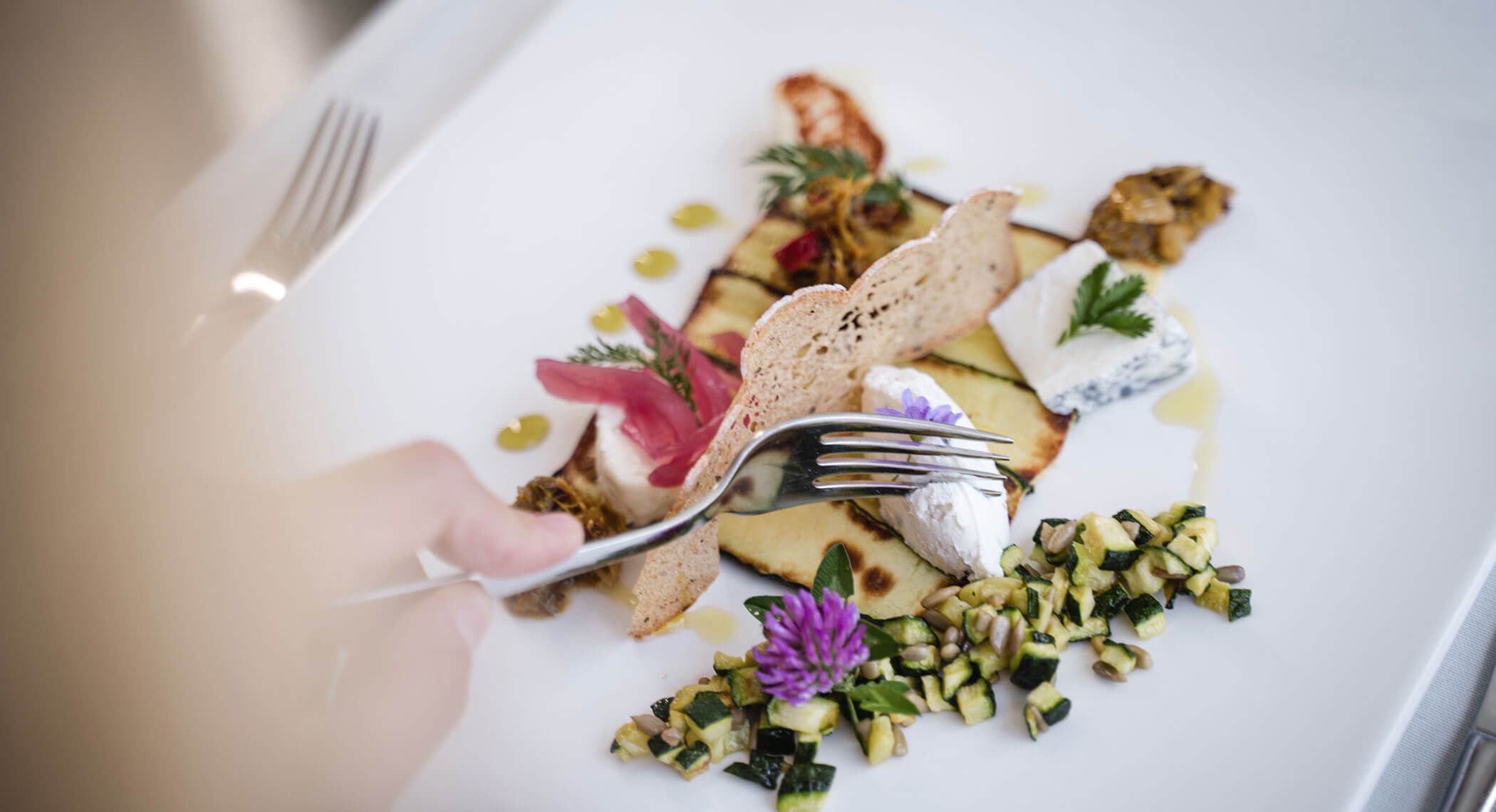 Eine Gabel, die Käse von einem Gourmet-Teller mit Gemüse, Kräutern und essbaren Blumen auf einem weißen Teller aufnimmt. - Hotel Moosmair