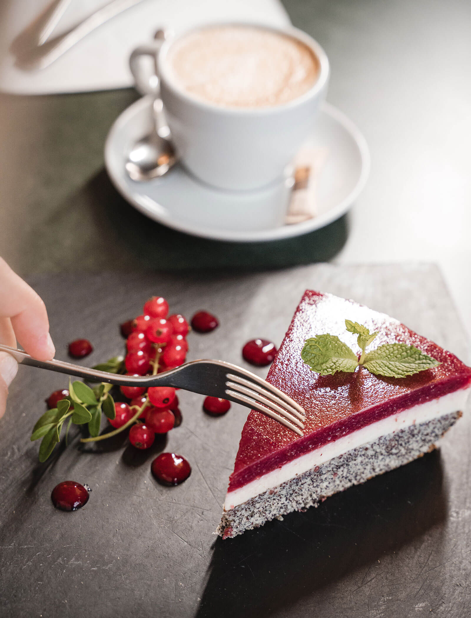 Eine Gabel schneidet in eine Beeren-Mousse-Torte mit Minze, roten Johannisbeeren und einer Tasse Kaffee im Hintergrund. - Hotel Moosmair