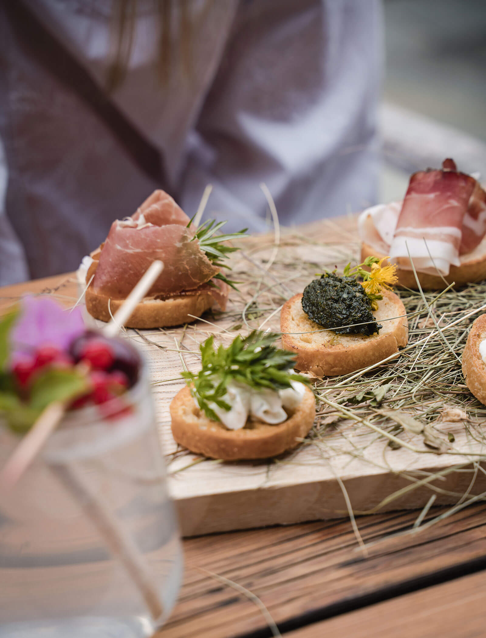 Ein Holzbrett mit Gourmet-Häppchen auf Brotscheiben, belegt mit Kräutern, Prosciutto und Aufstrich. - Hotel Moosmair