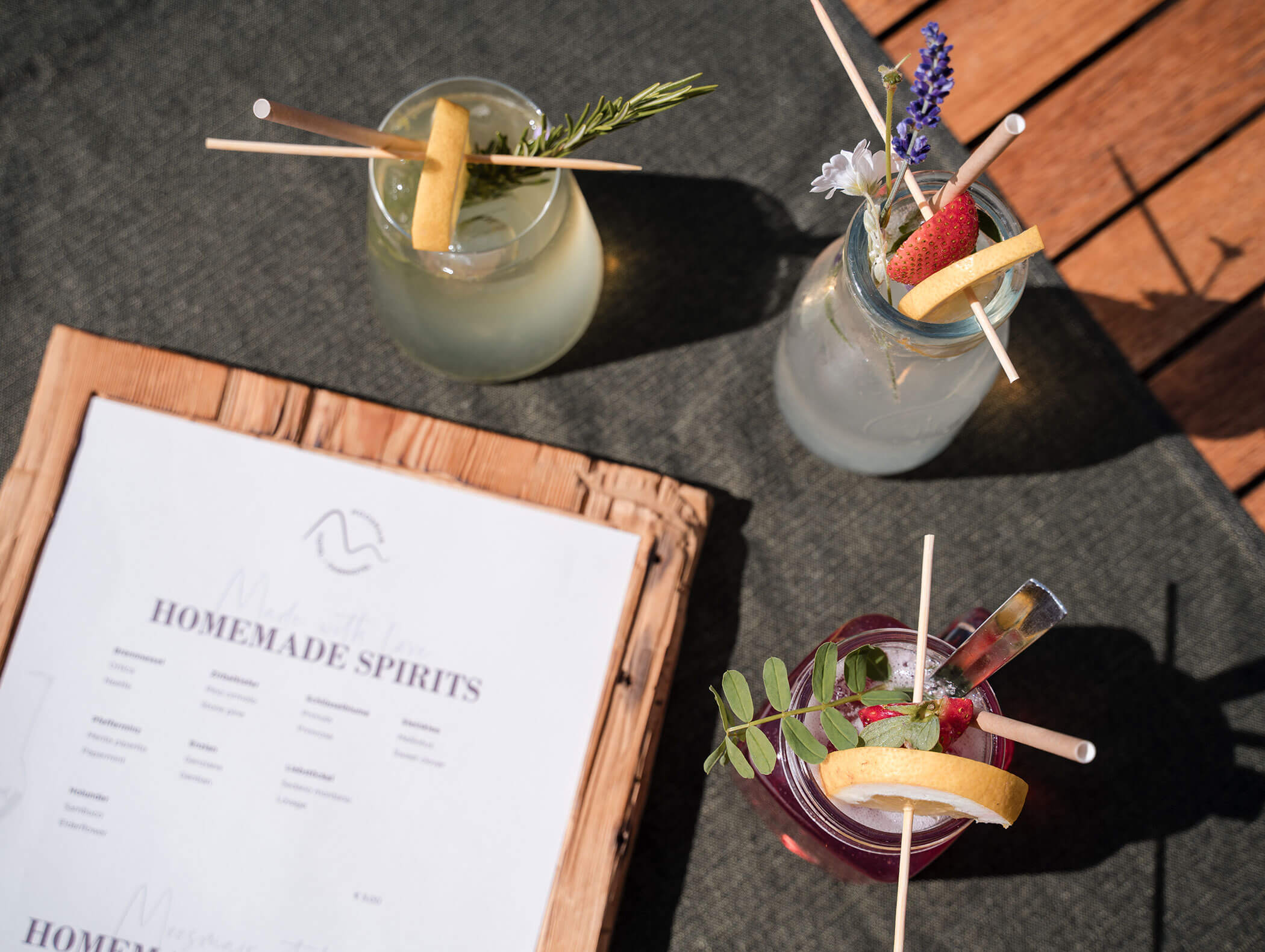 Drei bunte Cocktails mit Garnierungen neben einer hausgemachten Spirituosenkarte auf einem hölzernen Klemmbrett, im Freien. - Hotel Moosmair