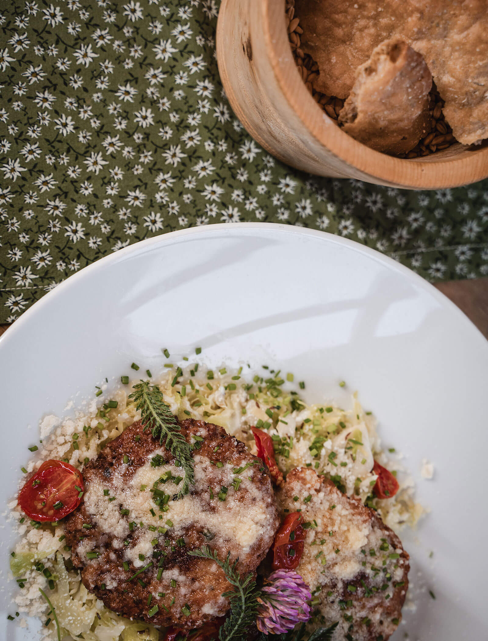 Zwei knusprige Patties auf einem weißen Teller mit geraspeltem Kohl, Tomaten, Kräutern und geriebenem Käse. Brot in einer Holzschale. - Hotel Moosmair