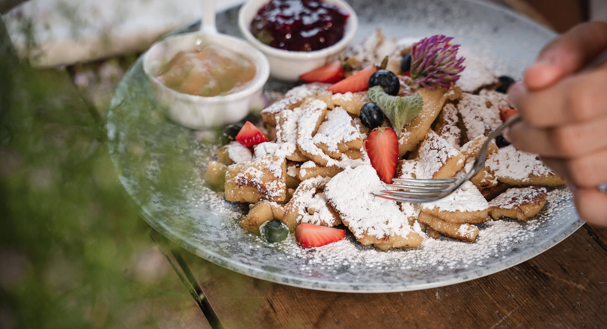 Ein Teller mit Puderzucker bestäubten Pfannkuchen mit Erdbeeren, Blaubeeren und Soße auf einem Holztisch. - Hotel Moosmair