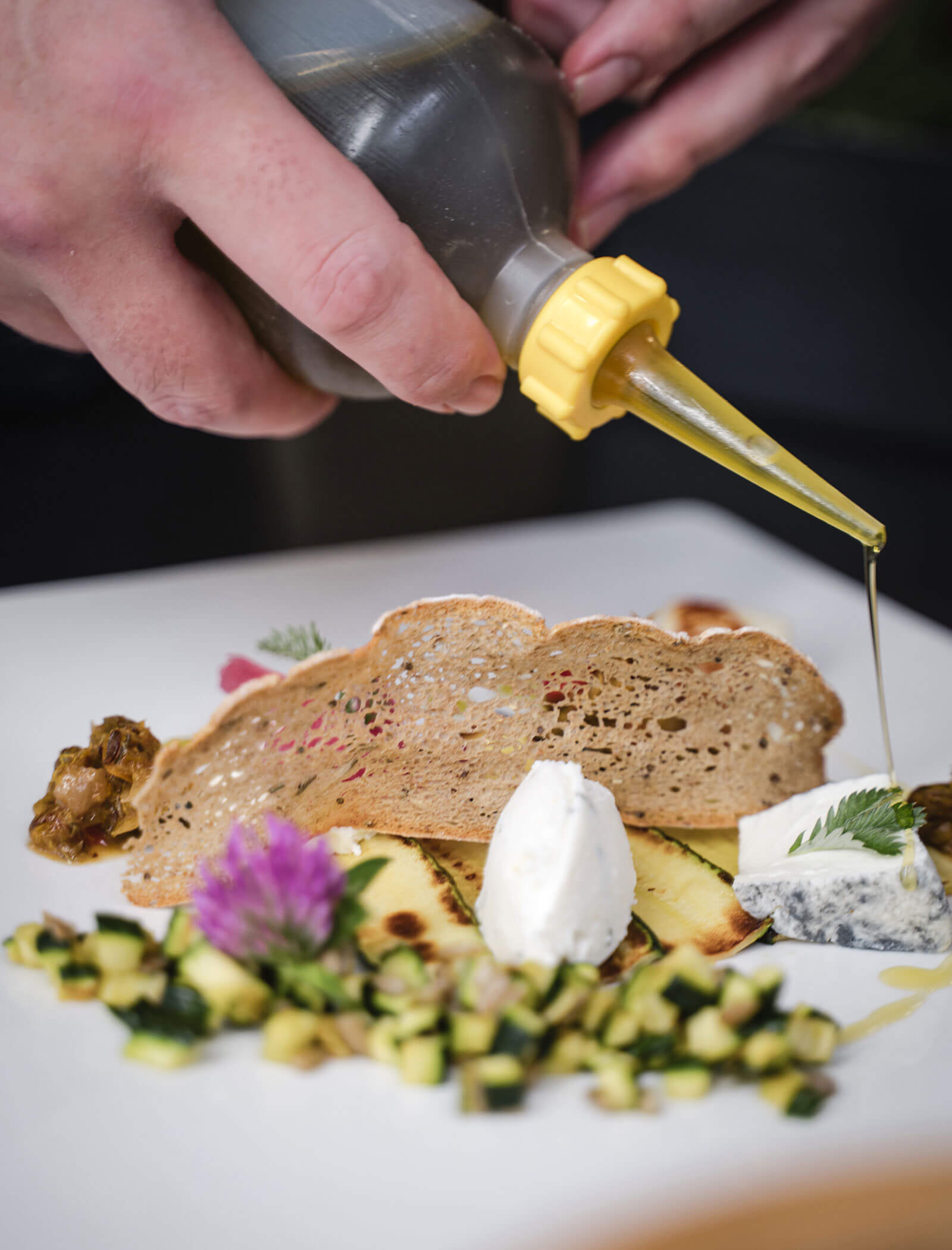 Eine Hand träufelt Öl über ein Gourmetgericht mit Brot, Käse, Kräutern und gewürfeltem Gemüse auf einem weißen Teller. - Hotel Moosmair