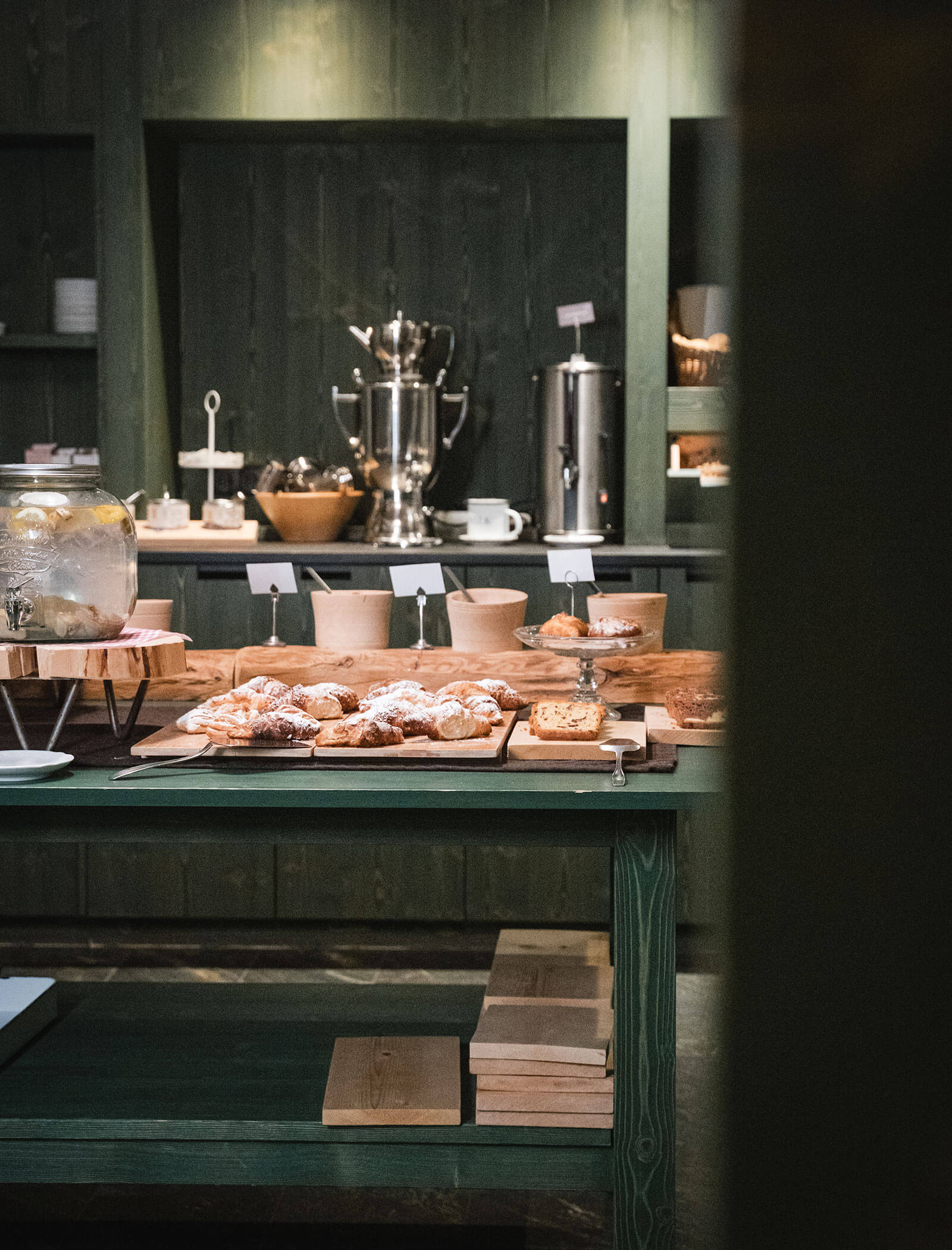 Holztisch mit verschiedenen Backwaren, Brot und Getränken in einem gemütlichen, rustikalen Selbstbedienungs-Frühstücksraum. - Hotel Moosmair