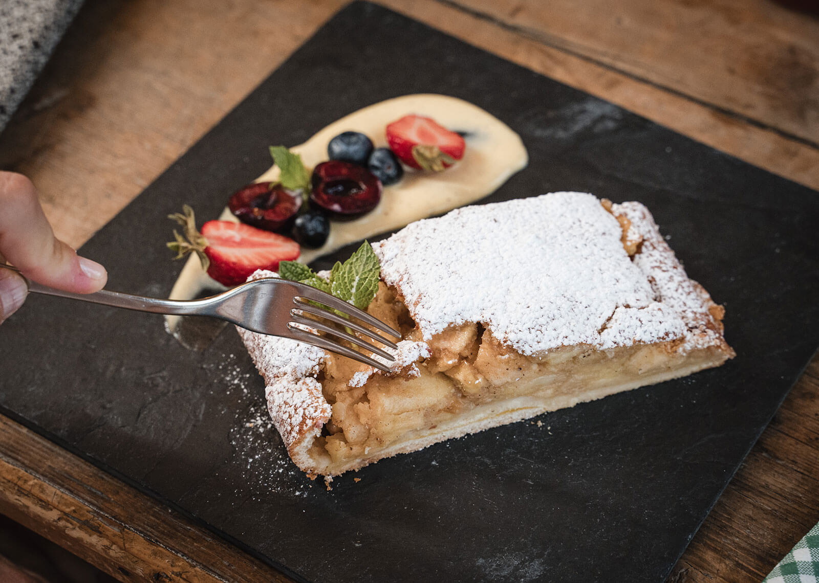 Eine Gabel, die in einen Apfelstrudel mit Puderzucker schneidet, der mit Sahne und frischen Beeren auf einem schwarzen Teller serviert wird. - Hotel Moosmair