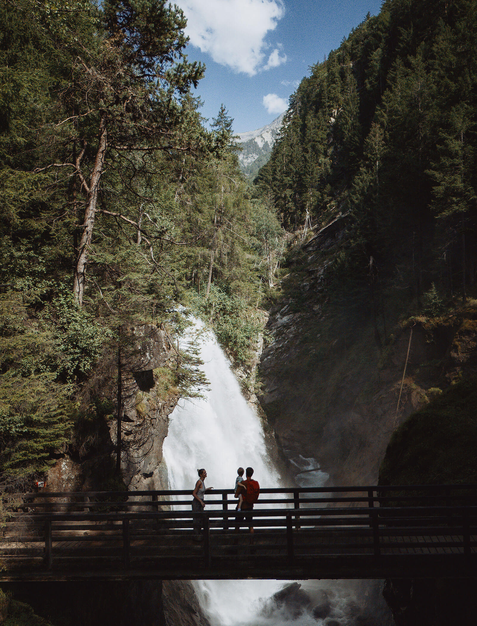 Drei Personen stehen auf einer Holzbrücke vor einem Wasserfall, der von hohen Bäumen und Bergen umgeben ist. - Hotel Moosmair