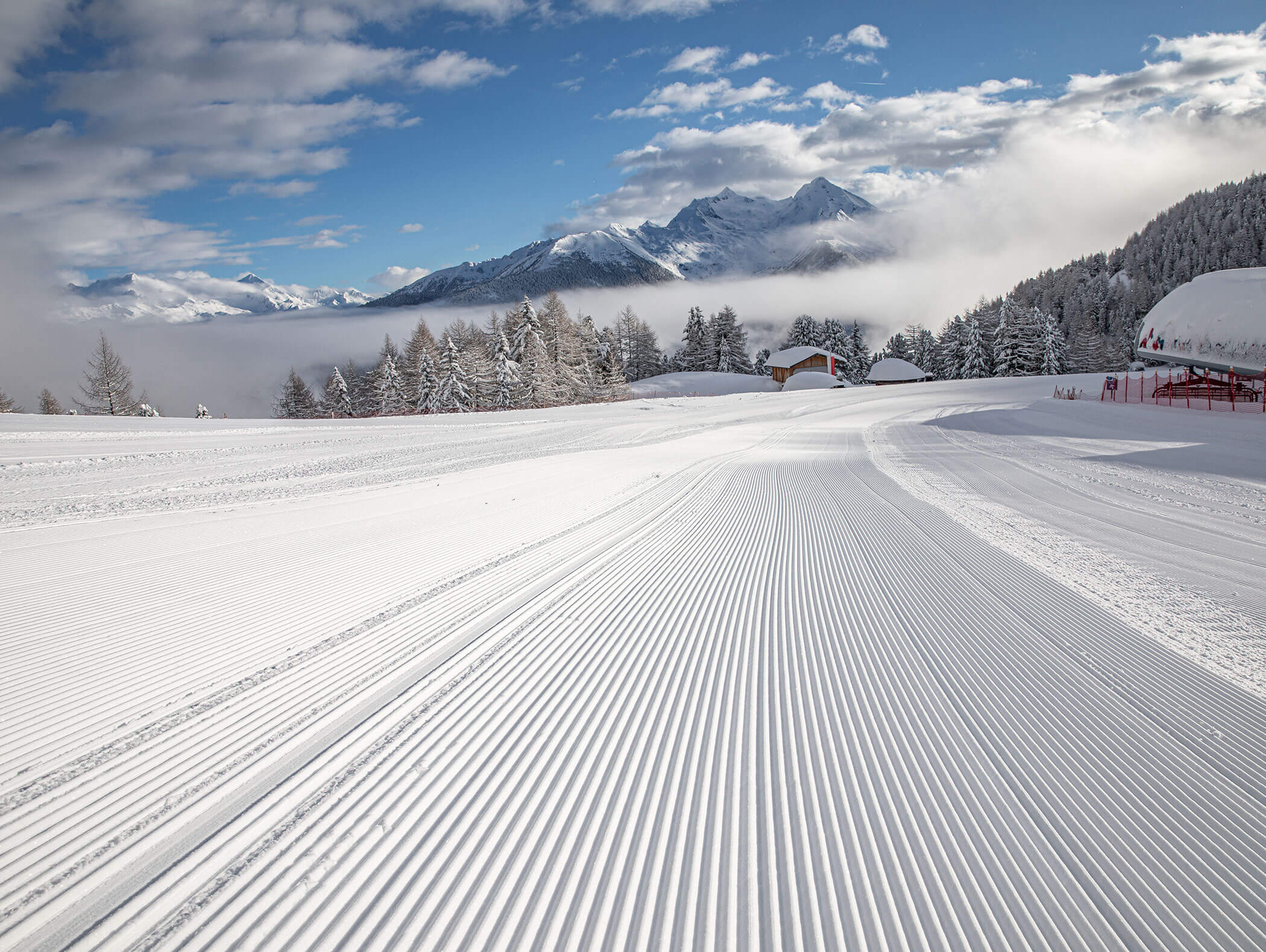 Frisch präparierte Skipiste mit schneebedeckten Bäumen und Bergen unter einem teilweise bewölkten blauen Himmel. - Hotel Moosmair