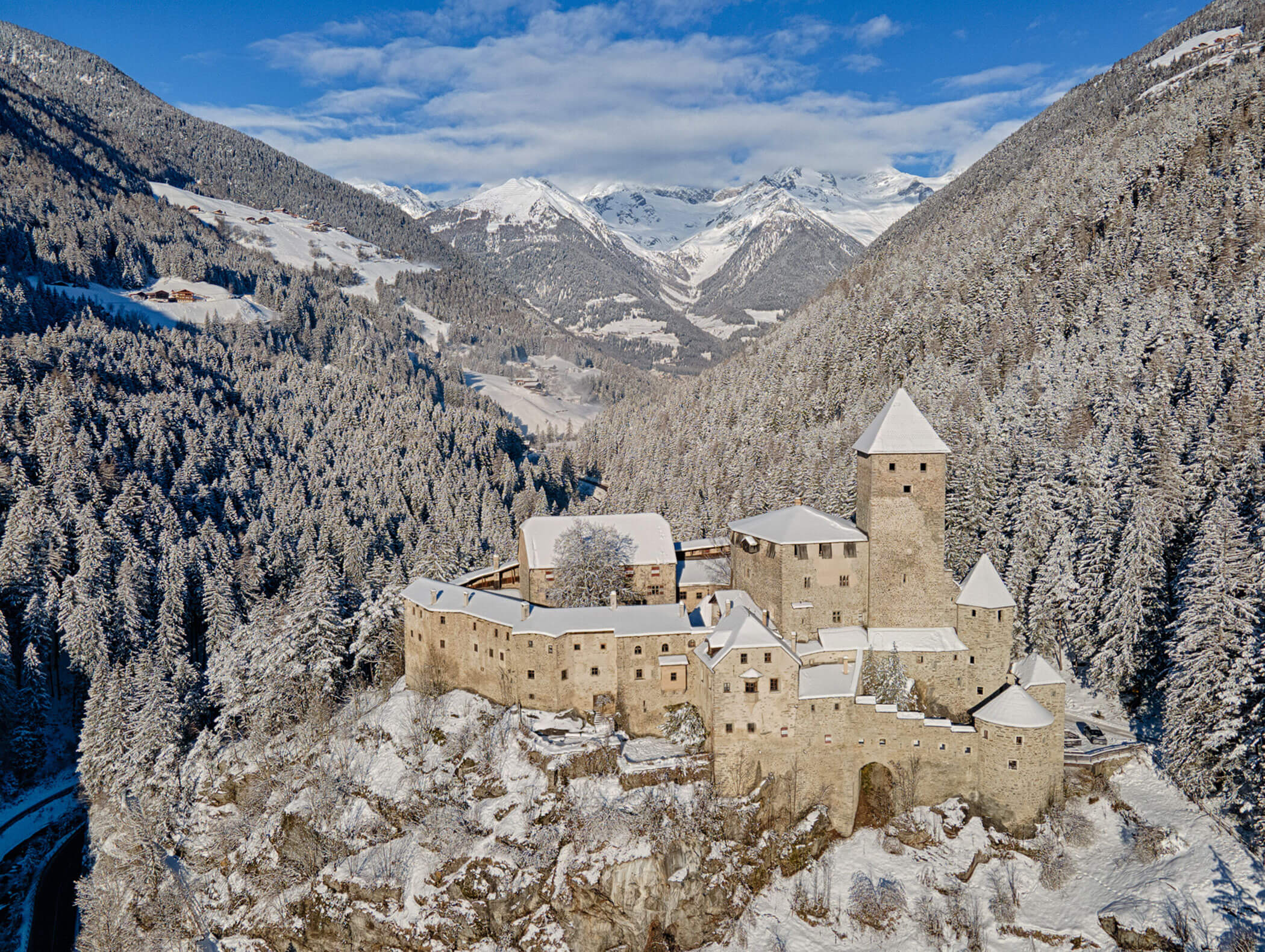 Ein schneebedecktes Schloss steht auf einem Hügel, umgeben von Kiefernwäldern und Bergen unter blauem Himmel. - Hotel Moosmair