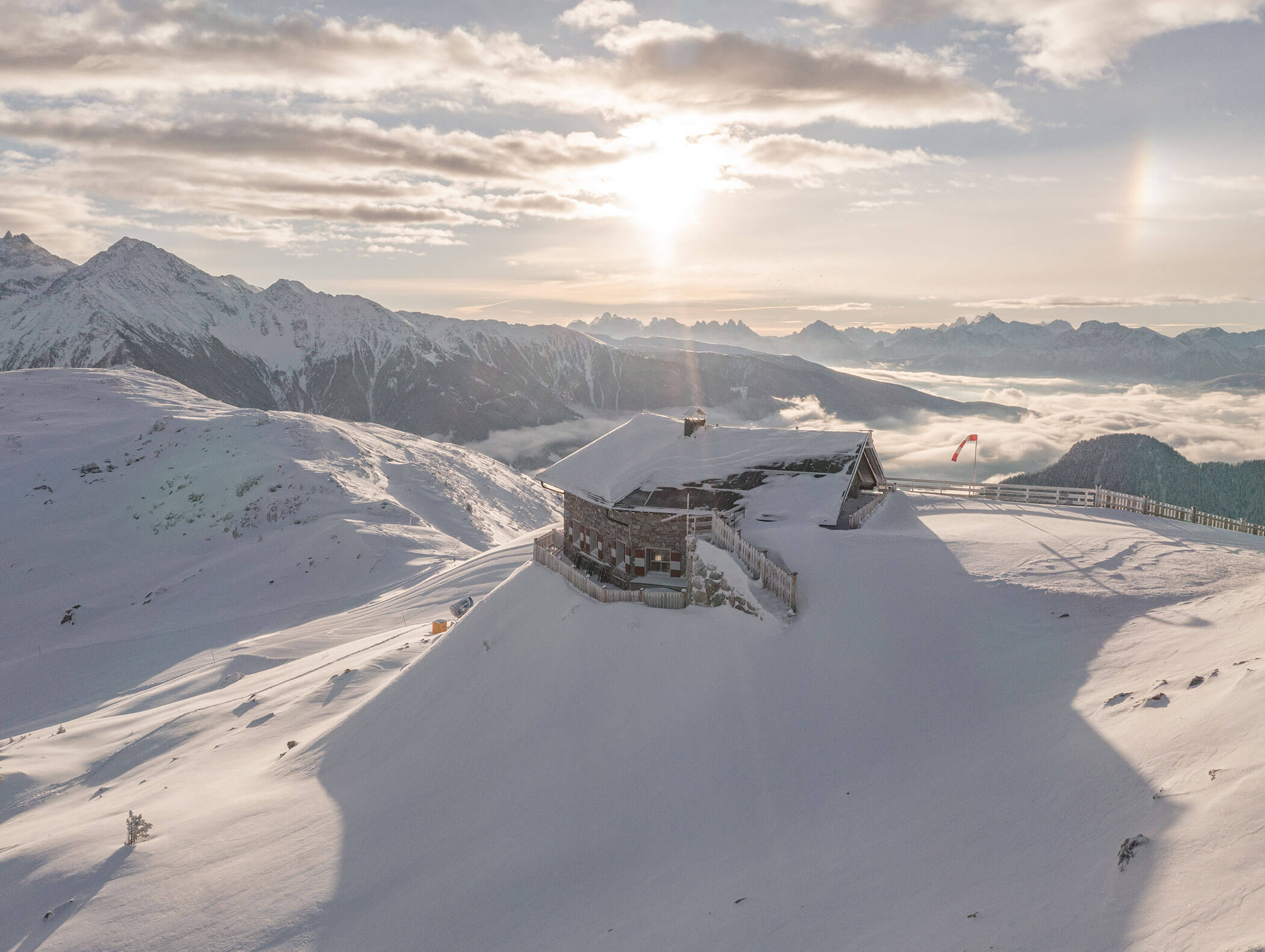 Eine verschneite Berghütte bei Sonnenaufgang, umgeben von schneebedeckten Gipfeln und Wolken im Tal darunter. - Hotel Moosmair