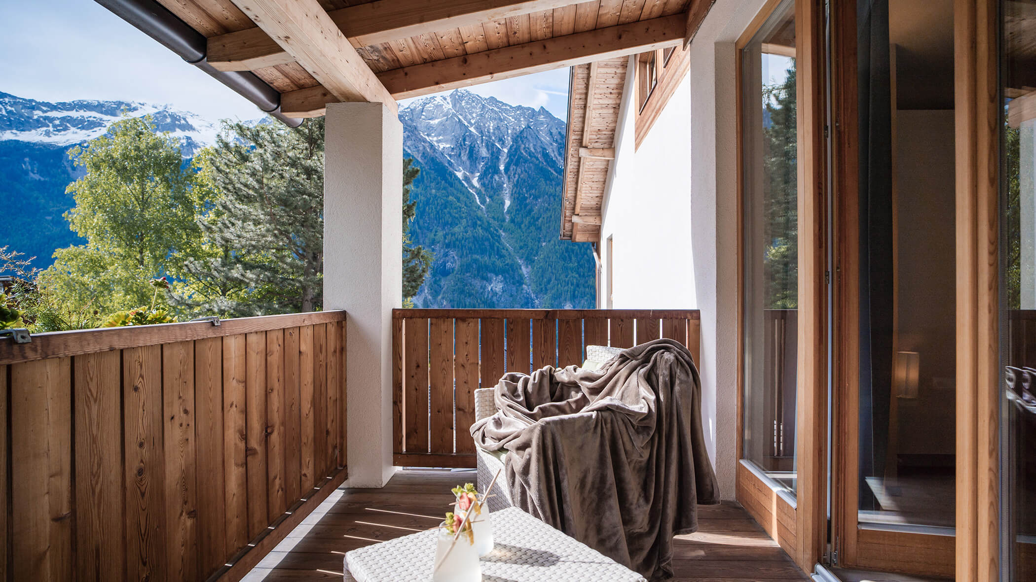Ein gemütlicher Bergbalkon mit Holzgeländer, einem Stuhl mit einer Decke und beeindruckenden Schneegipfeln im Hintergrund. - Hotel Moosmair