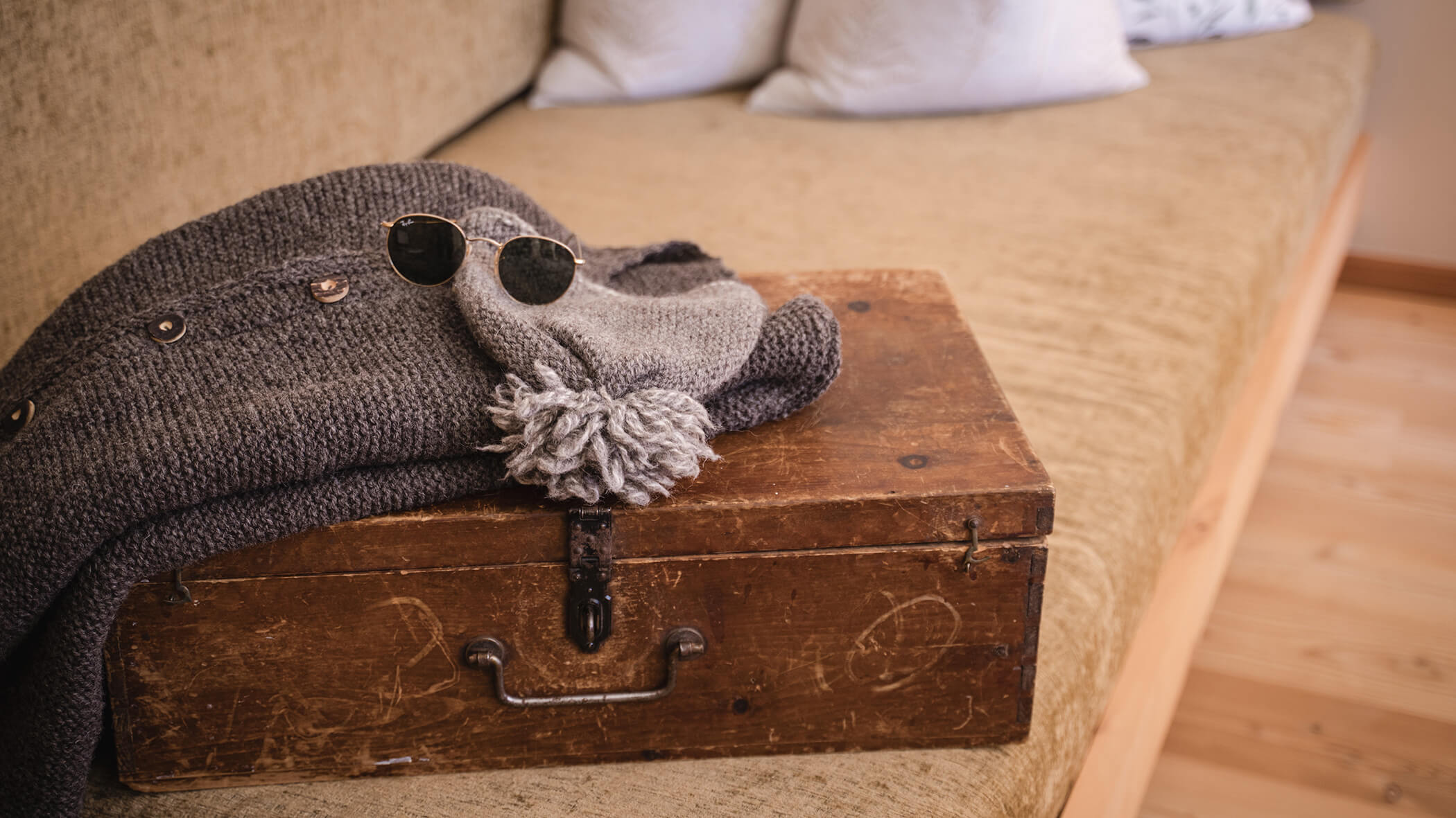 Ein Strickpullover, eine Mütze und eine runde Sonnenbrille liegen auf einer alten Holztruhe auf einem beigen Sofa. - Hotel Moosmair
