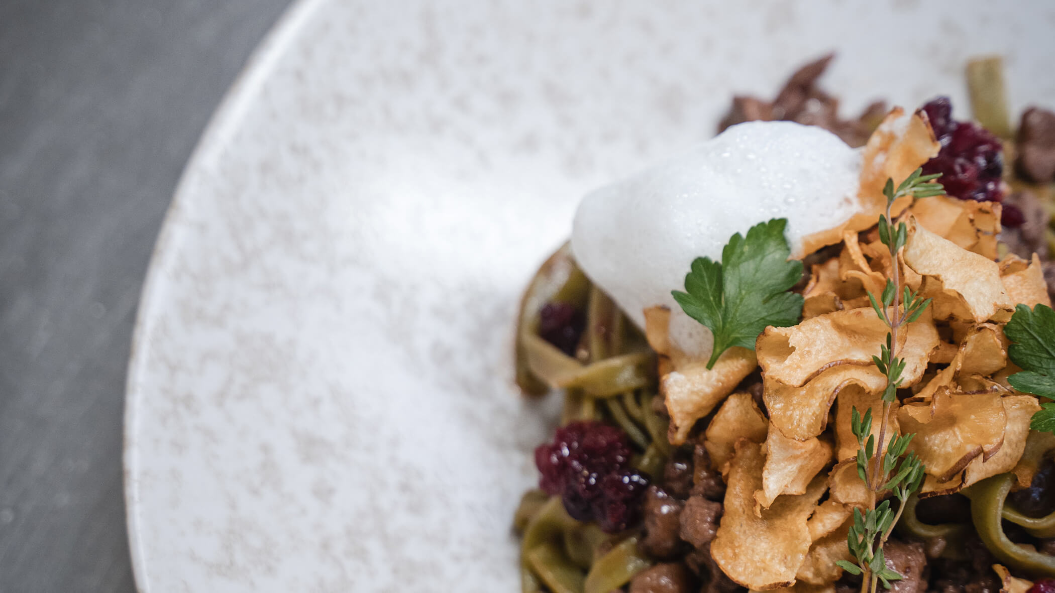 Nahaufnahme von Gourmet-Nudeln mit knusprigen Chips, Schaum, Kräutern und einer Beerengarnitur auf einem weißen Teller. - Hotel Moosmair
