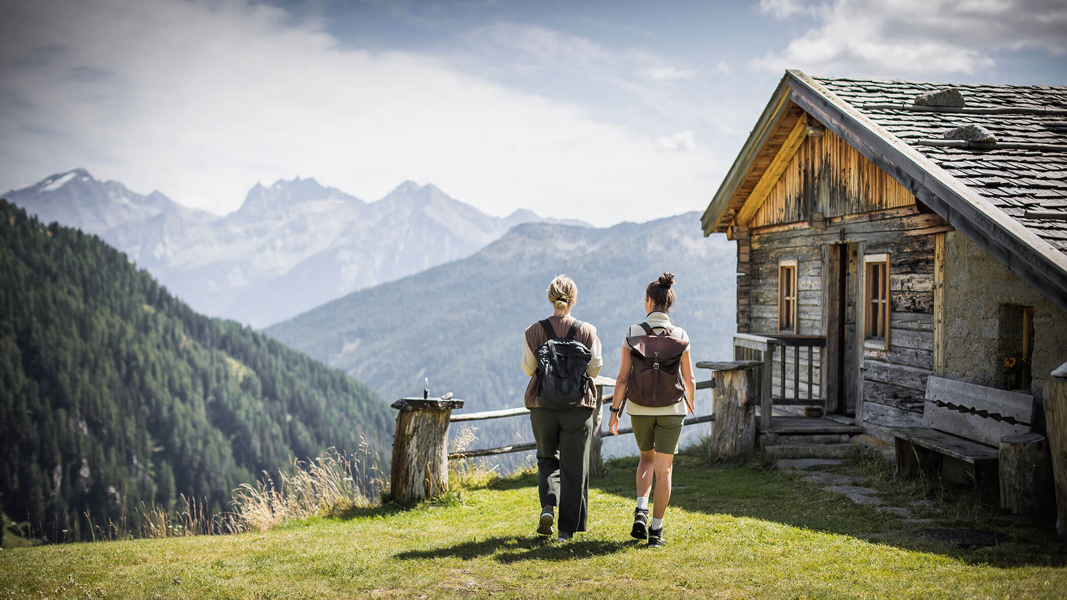 Zwei Personen mit Rucksäcken gehen an einem sonnigen Tag auf eine rustikale Holzhütte in den Bergen zu. - Hotel Moosmair