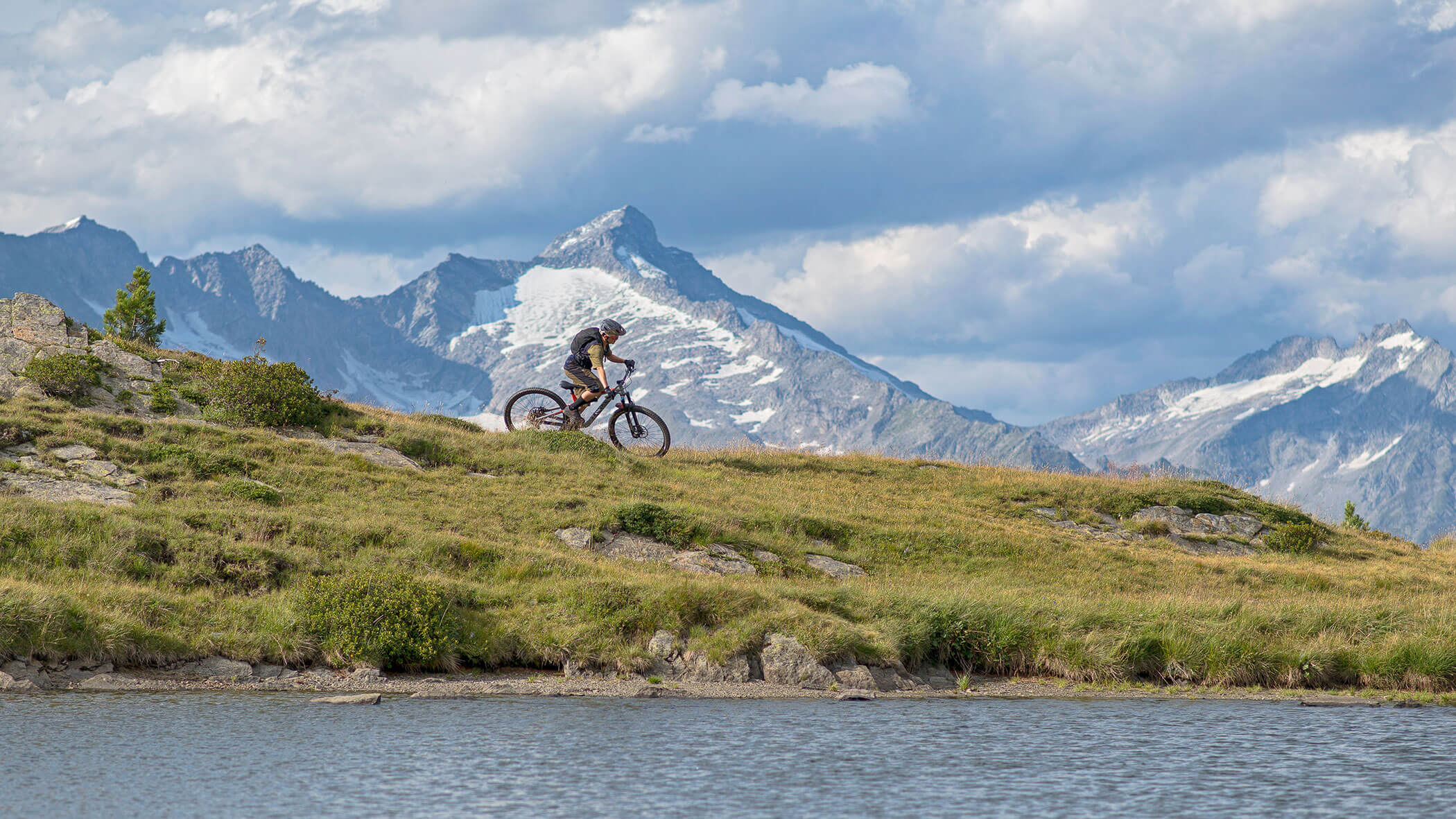 Ein Radfahrer fährt auf einem grasbewachsenen Hügel am Wasser entlang, mit schneebedeckten Bergen im Hintergrund unter einem bewölkten Himmel. - Hotel Moosmair