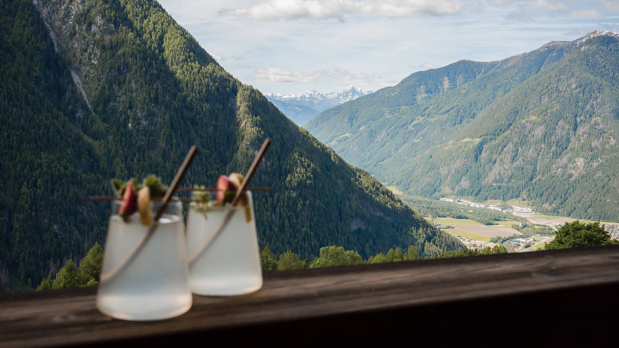 Zwei Cocktails auf einem Holzgeländer mit Blick auf grüne Berge und ein Tal unter einem teilweise bewölkten Himmel. - Hotel Moosmair