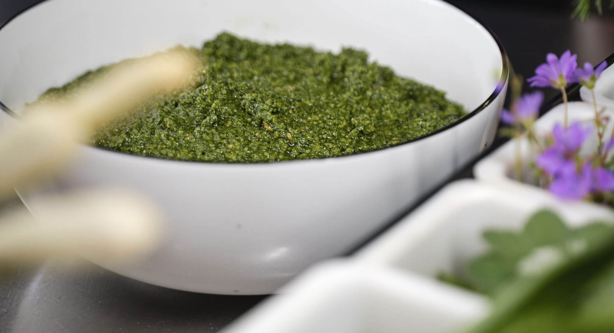 Eine weiße Schale mit grüner Pestosauce, daneben Kräuter und lila Blumen. - Hotel Moosmair