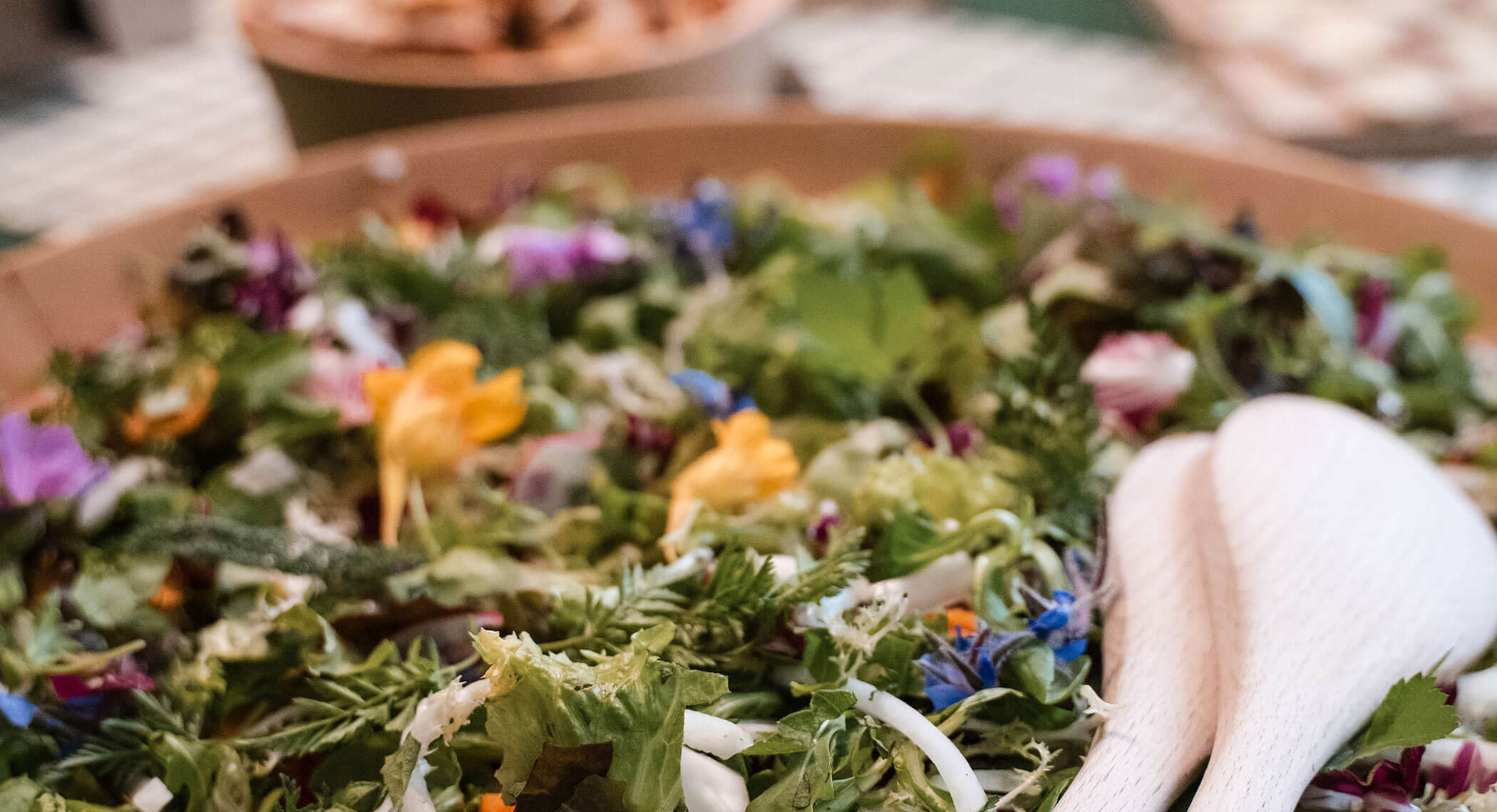 Ein bunter gemischter Salat mit essbaren Blumen in einer Schüssel, mit einem hölzernen Salatbesteck an der Seite. - Hotel Moosmair
