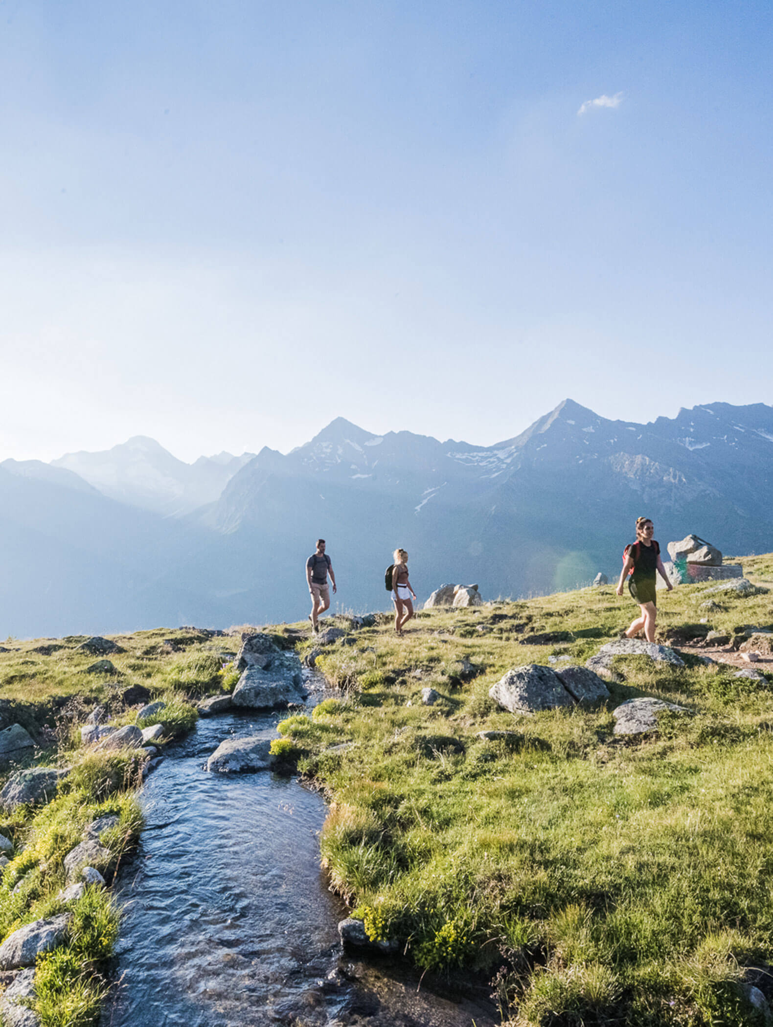 Drei Personen wandern entlang eines Baches in einer grasbewachsenen Berglandschaft mit Gipfeln im Hintergrund unter einem klaren Himmel. - Hotel Moosmair