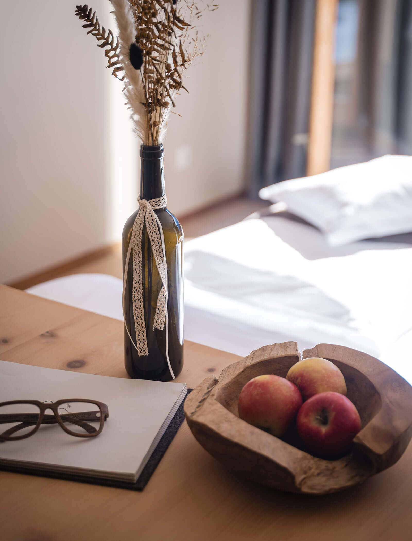 Ein Holztisch mit einem Notizbuch, Gläsern, einer Vase mit Trockenblumen und einer Holzschale mit Äpfeln. - Hotel Moosmair