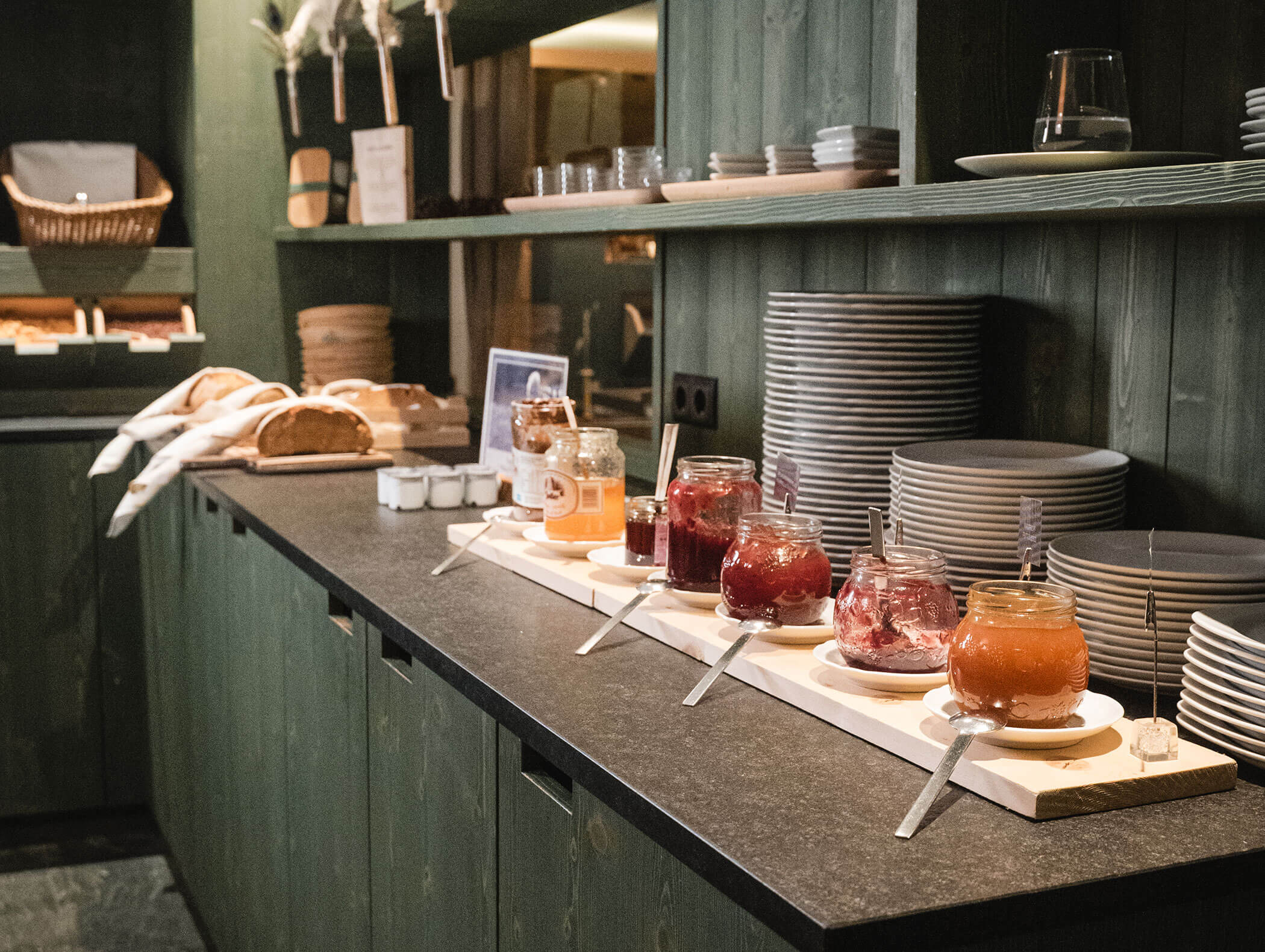 Verschiedene Gläser mit Marmelade, Brot und Teller sind auf einer dunklen Arbeitsplatte in einer rustikalen Küche angeordnet. - Hotel Moosmair