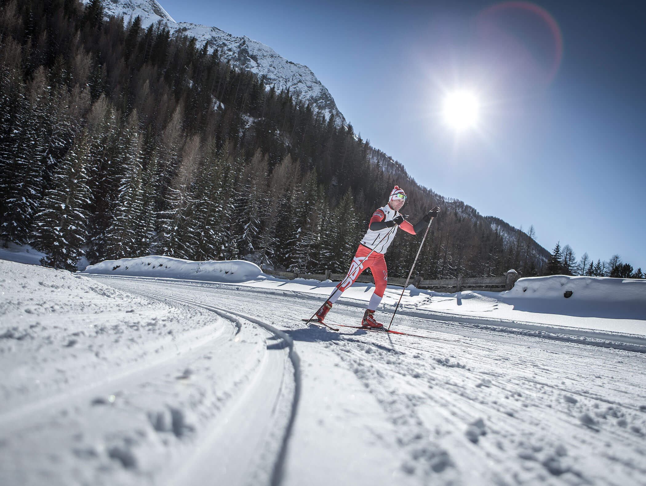 Eine Person beim Skilanglauf auf einer verschneiten Loipe mit Bergen und Bäumen bei strahlendem Sonnenschein. - Hotel Moosmair