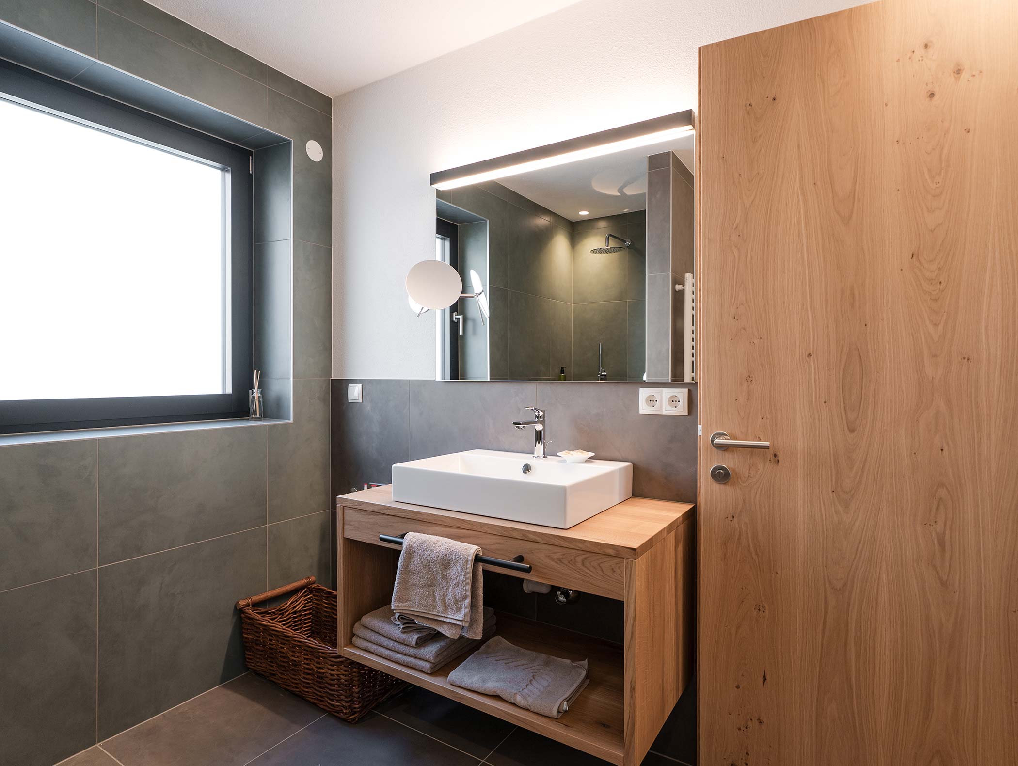Modernes Badezimmer mit einem hölzernen Waschtisch, einem quadratischen Waschbecken, einem großen Spiegel und ordentlich gefalteten Handtüchern auf einem Regal. - Hotel Moosmair