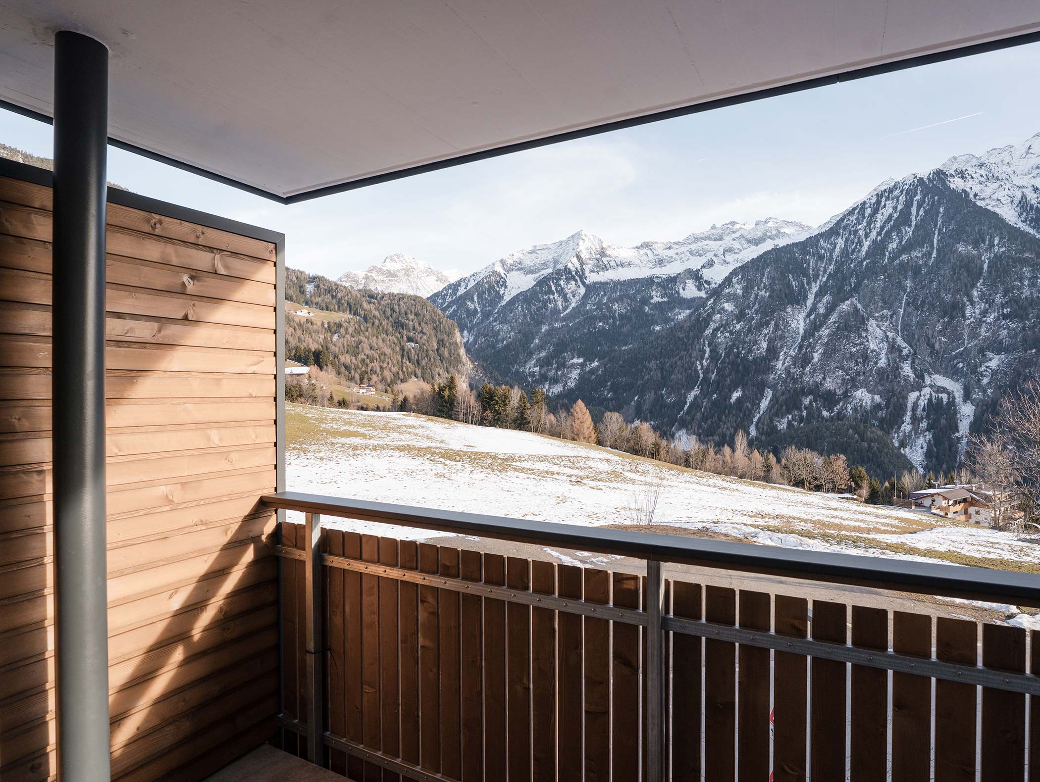 Blick von einem Holzbalkon auf schneebedeckte Berge und eine grasbewachsene Wiese bei klarem Himmel. - Hotel Moosmair