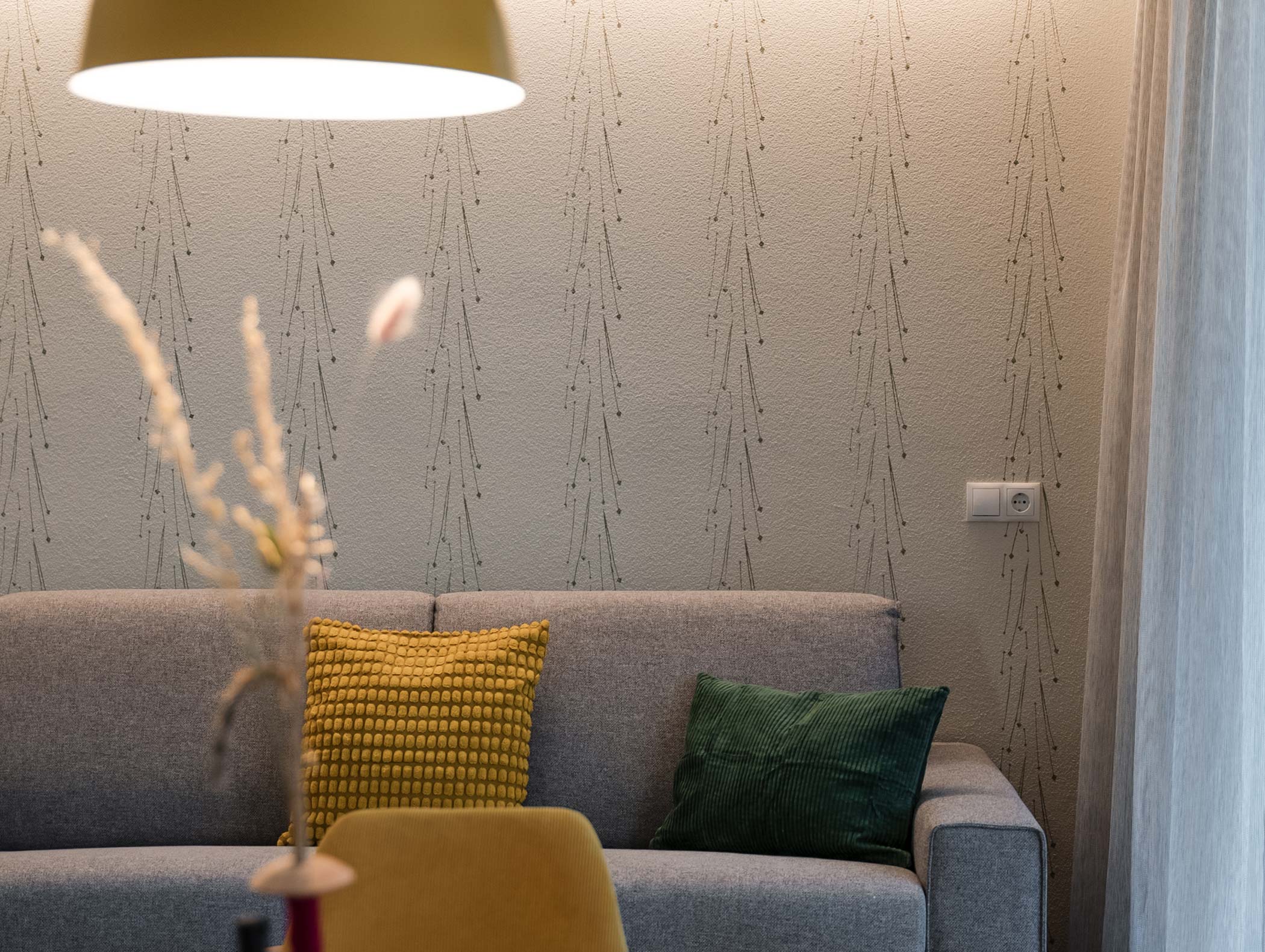 Graues Sofa mit gelben und grünen Kissen, Lampe darüber und gemusterter Tapete in einem gemütlichen Wohnzimmer. - Hotel Moosmair