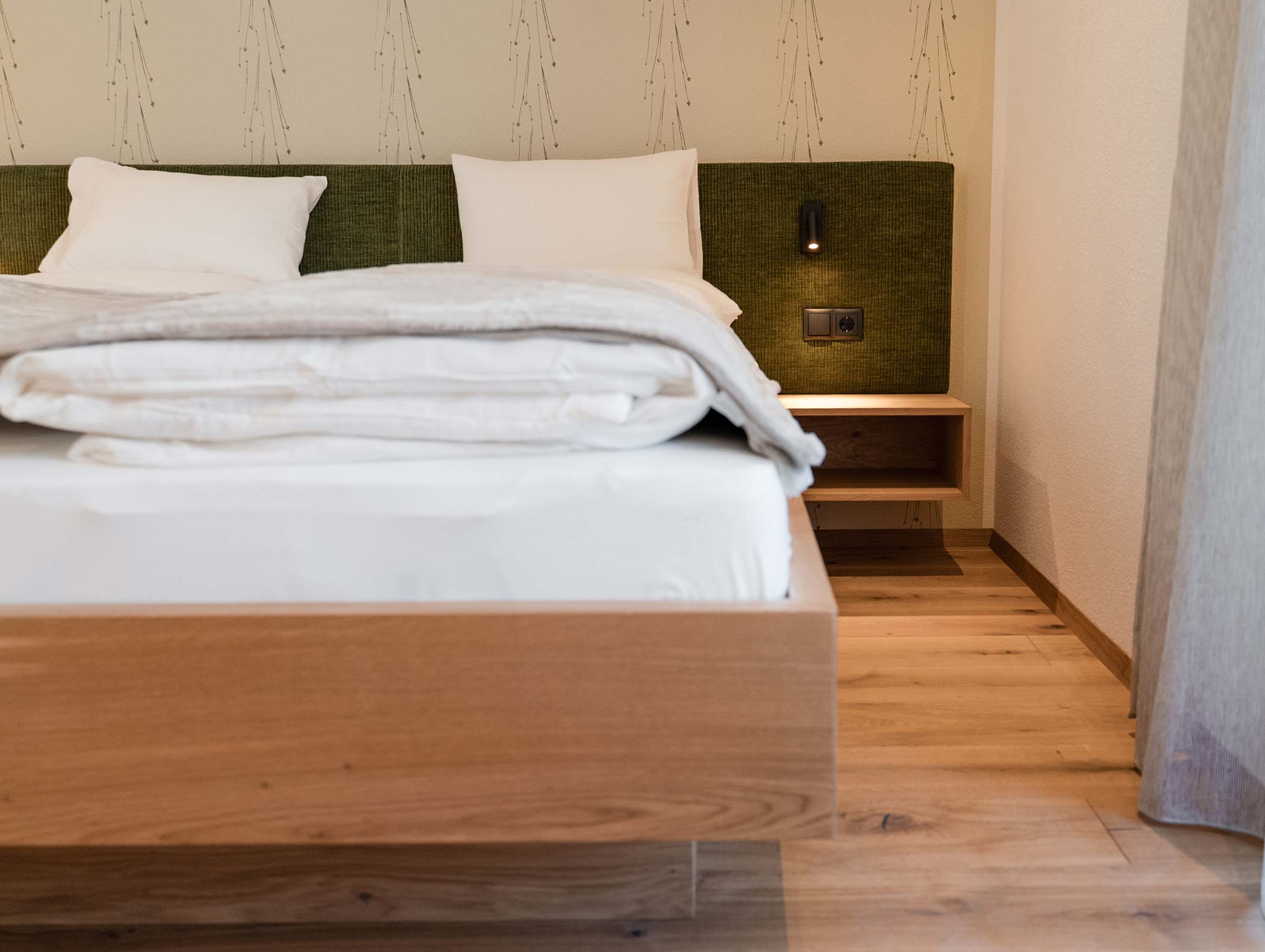 Minimalistisches Schlafzimmer mit sauber gemachtem Bett, grünem Kopfteil, Holzboden und Nachttisch mit Wandleuchte. - Hotel Moosmair