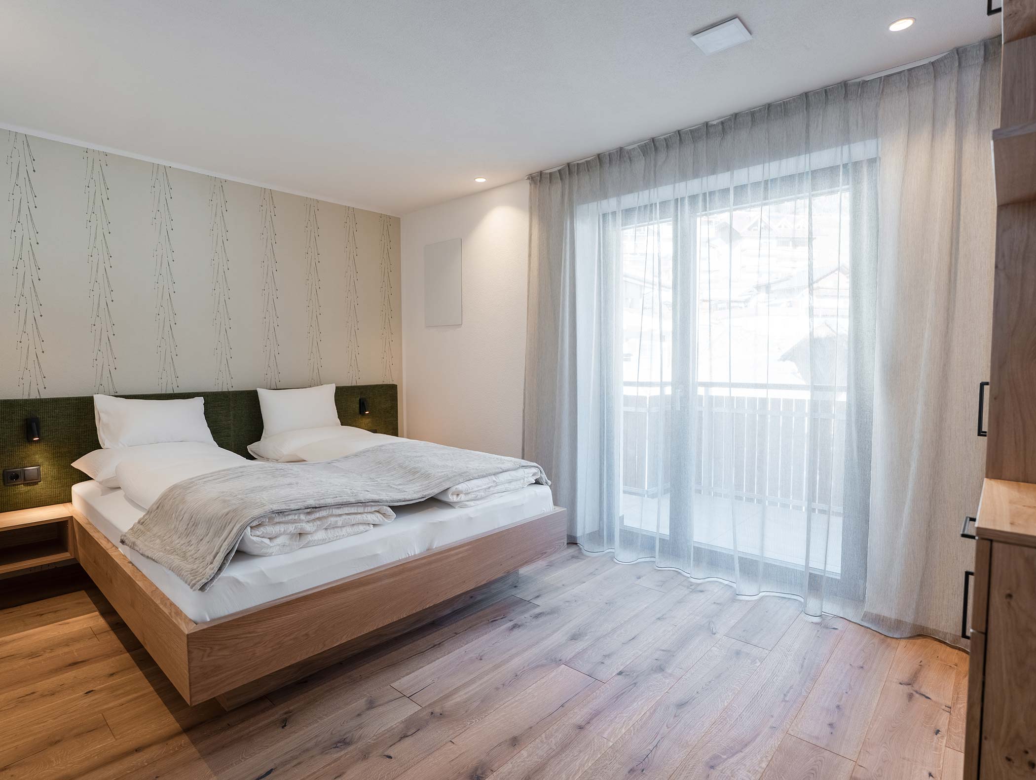 Modernes Schlafzimmer mit Doppelbett, hellem Holzboden und einem großen Fenster mit Vorhängen, die das Tageslicht hereinlassen. - Hotel Moosmair