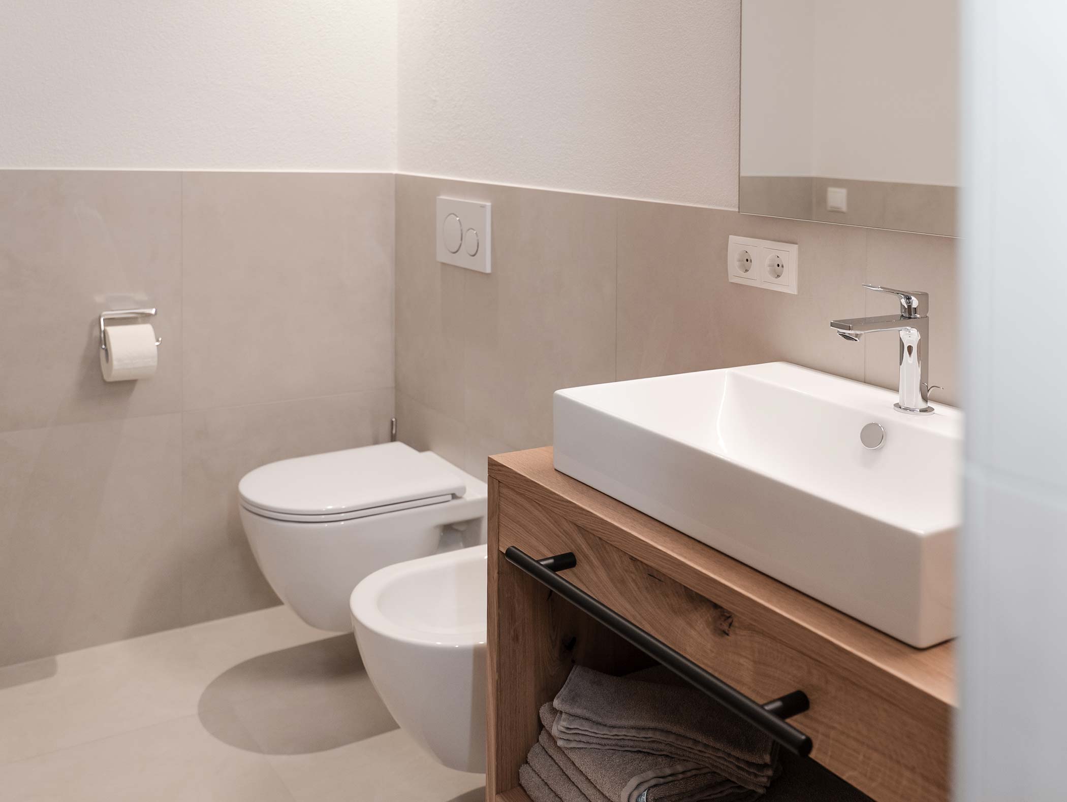 Modernes Badezimmer mit wandmontierter Toilette, Bidet, Waschbecken und hölzernem Waschtisch mit Handtüchern darunter. - Hotel Moosmair