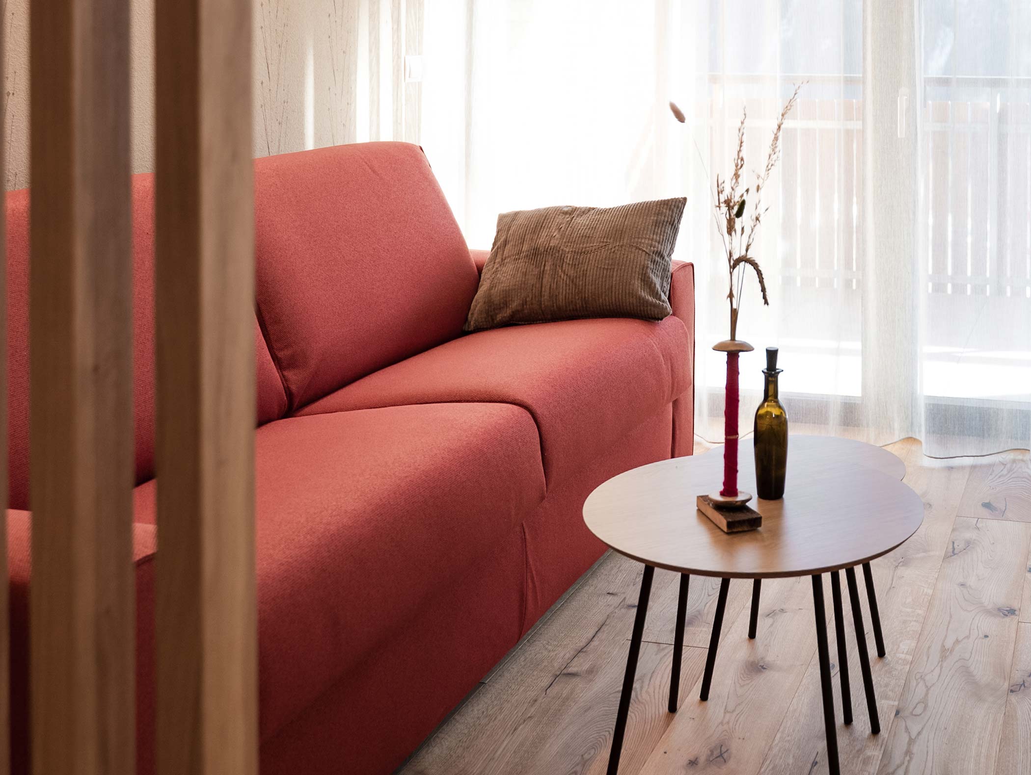 Ein modernes Wohnzimmer mit einem roten Sofa, einem Kissen und einem runden Couchtisch mit Dekoration an einem sonnigen Fenster. - Hotel Moosmair