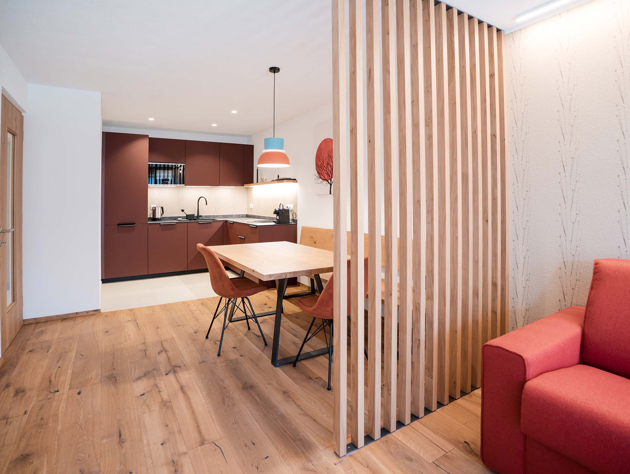 Moderne Küche und Essbereich mit Holzfußboden, roten Stühlen und einer Trennwand aus vertikalen Holzlatten. - Hotel Moosmair