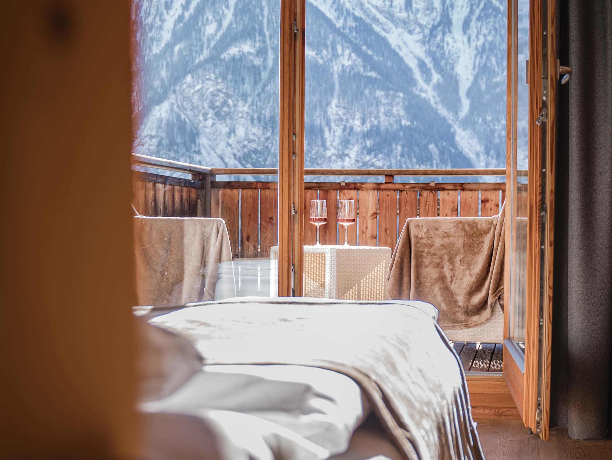 Ein gemütliches Schlafzimmer öffnet sich zu einem Balkon mit zwei Weingläsern und Bergblick im Hintergrund. - Hotel Moosmair